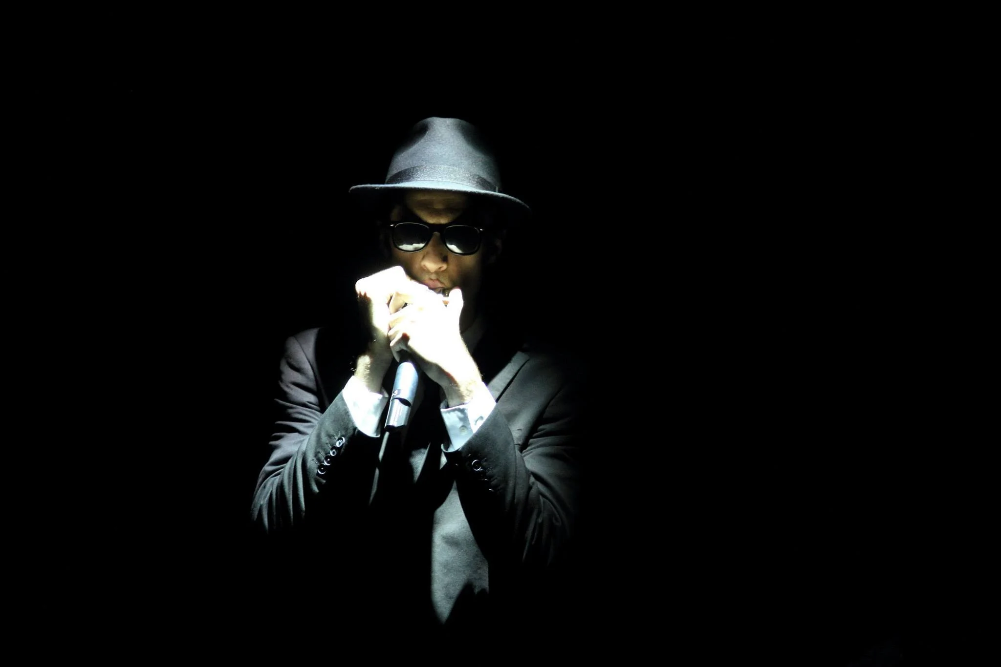 A person in a dark suit, white shirt, and glasses, wearing a fedora hat, is in a spotlight with their face partially lit as they hold a microphone and harmonica close to their mouth.