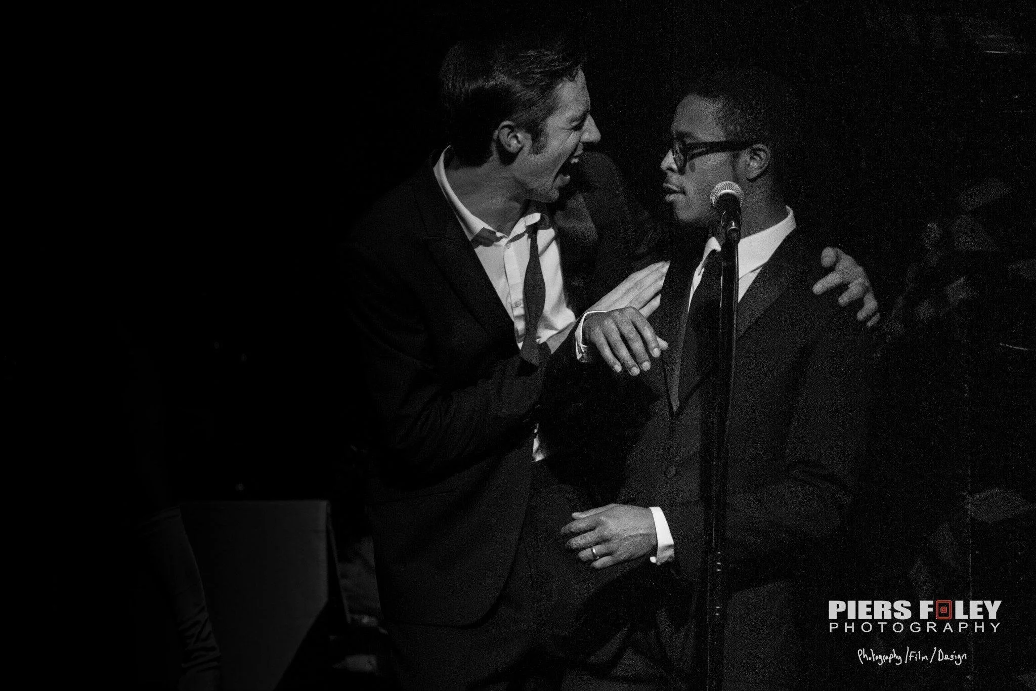 Two men dressed in suits are singing into a microphone while standing close together on a dark stage, with one man's hand on the other's shoulder.