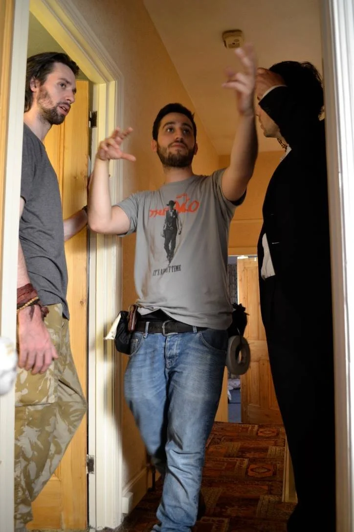 Three people in a hallway, engaged in conversation. One man in the center gestures with his hands, another man on the left in camouflage pants and a gray shirt, and a man on the right with curly hair and black attire.