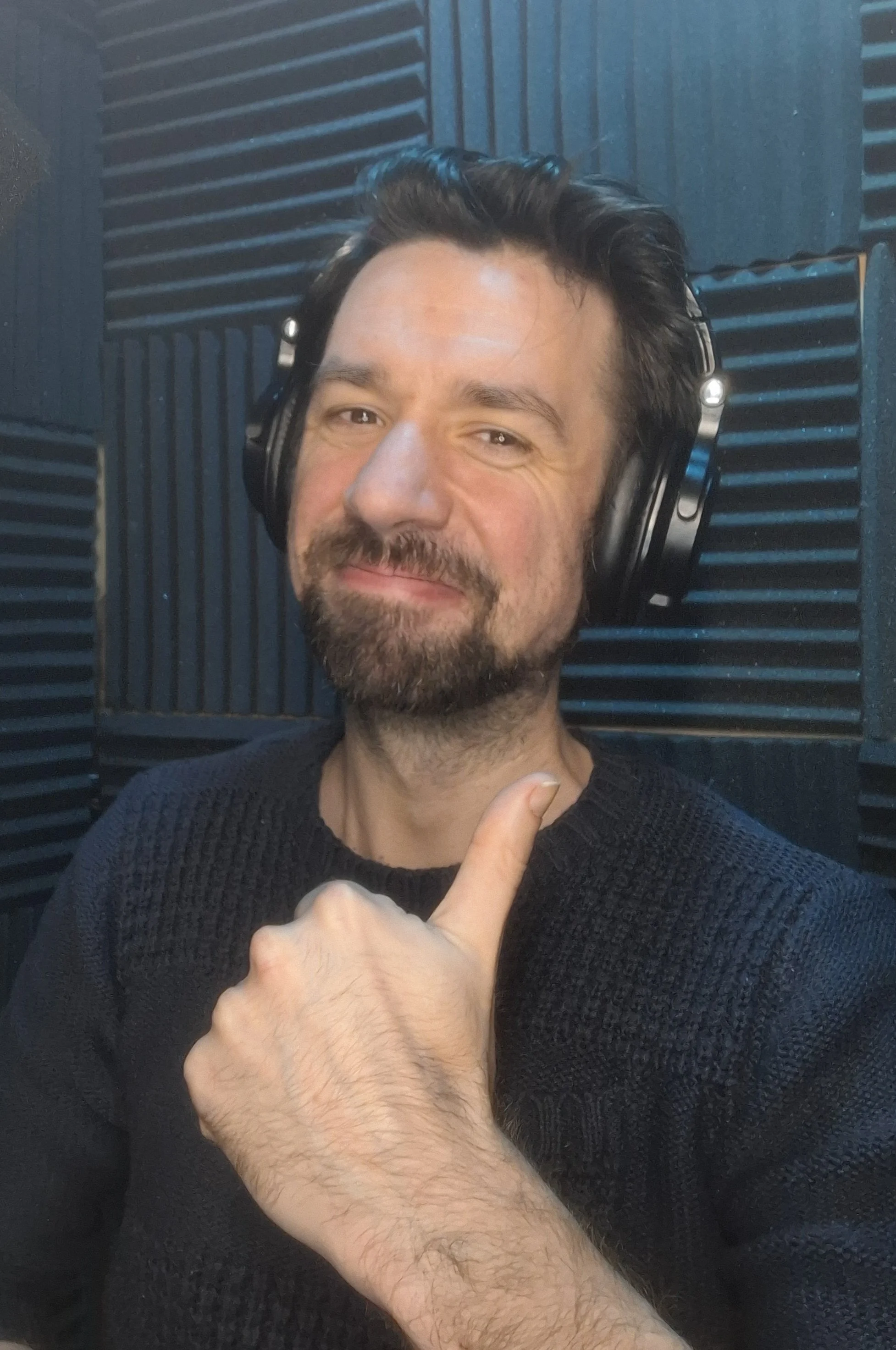 A man with dark hair, a beard, and light skin wearing a black sweater, smiling with a slightly squinting expression, in a recording studio. An arm with hand, pointing upward with the thumb, is visible in the foreground.