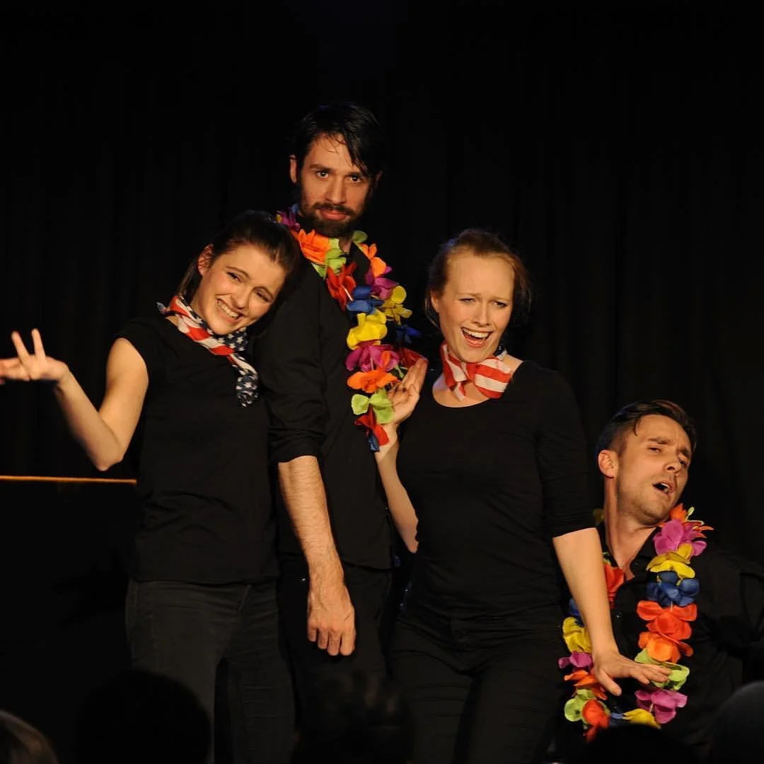 Four actors on stage dressed in black, wearing colorful leis and scarves, appearing to perform a comedic scene with various facial expressions.