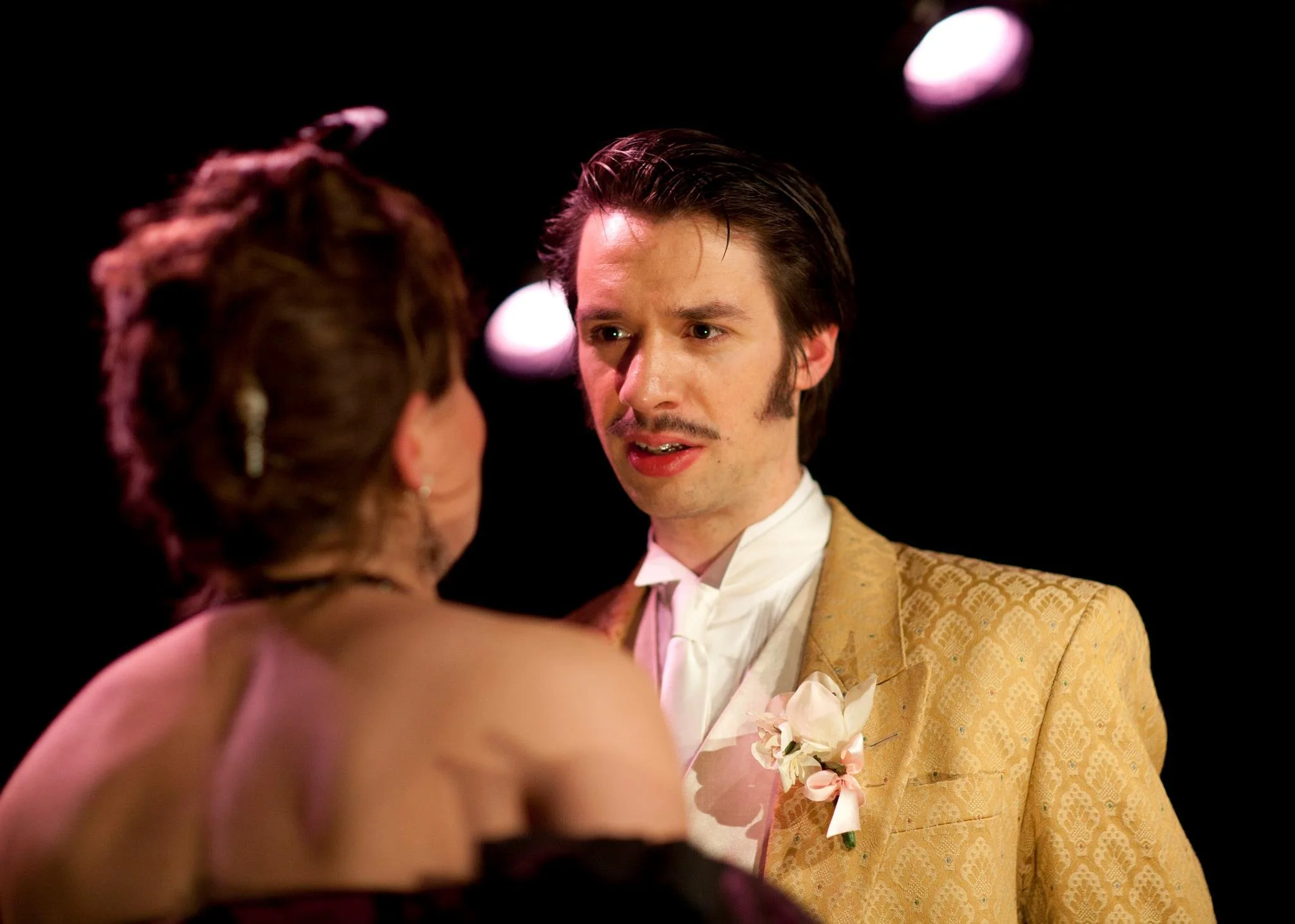 A man and woman stand closely, facing each other, in a dark background with some light spots. The man wears a gold patterned suit and white shirt, and the woman has styled hair and a dress.