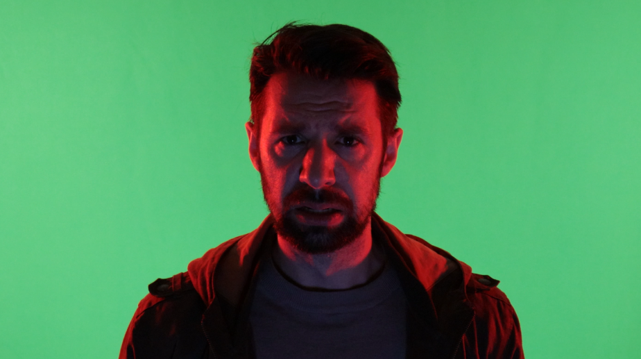 A man with a beard and short hair looking directly at the camera, illuminated by red and green lighting.