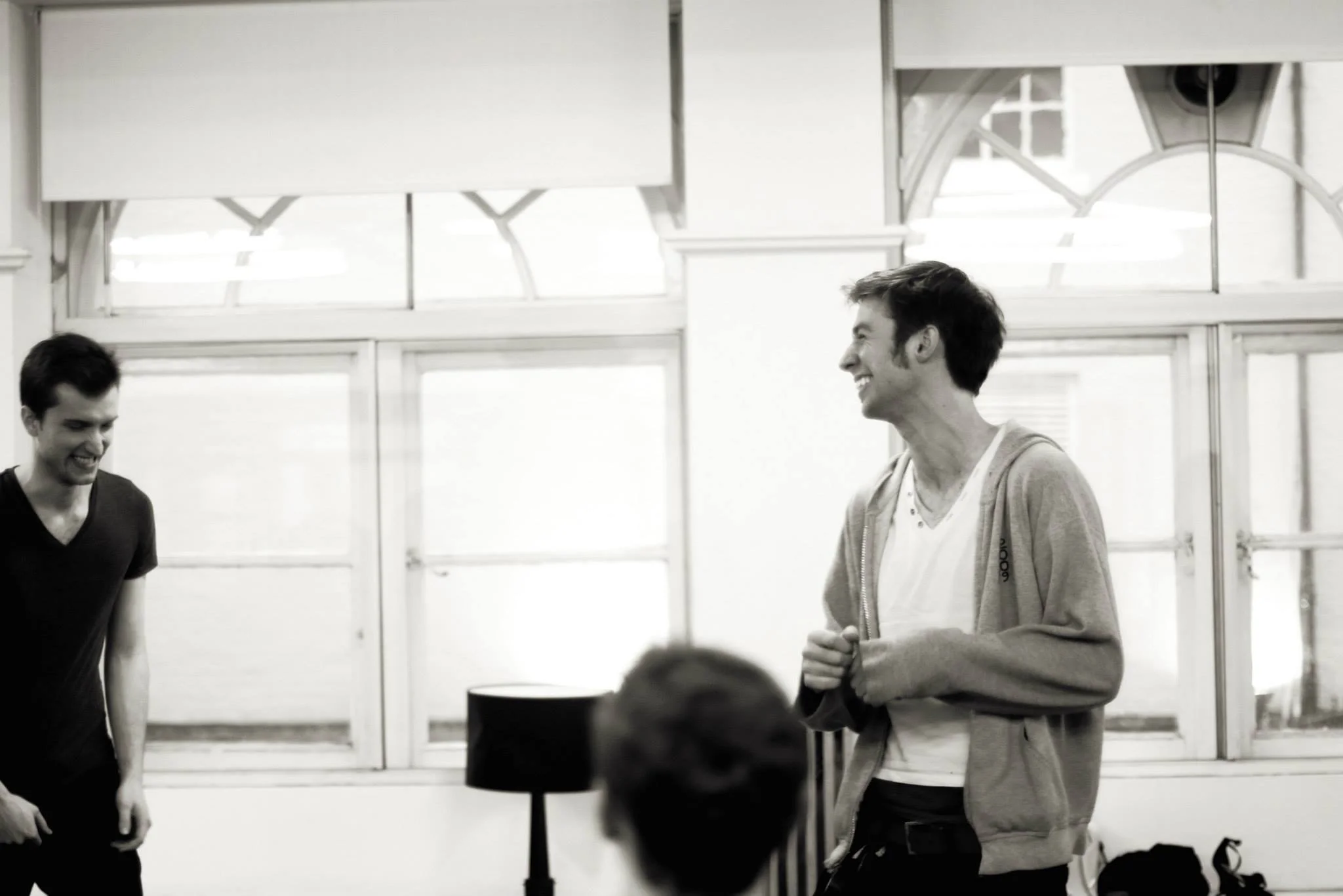 Two men smiling and engagement in conversation in a room with large windows and blinds, black and white photograph.