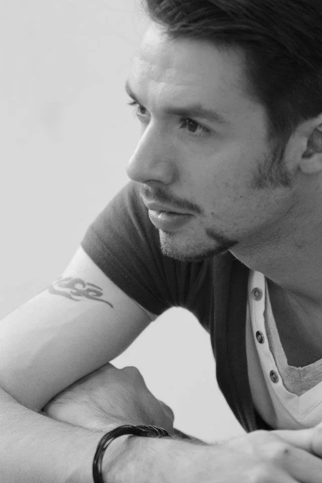 A young man with tattoos on his arm is looking to the side, sitting with his arms crossed, wearing a T-shirt and a bracelet.