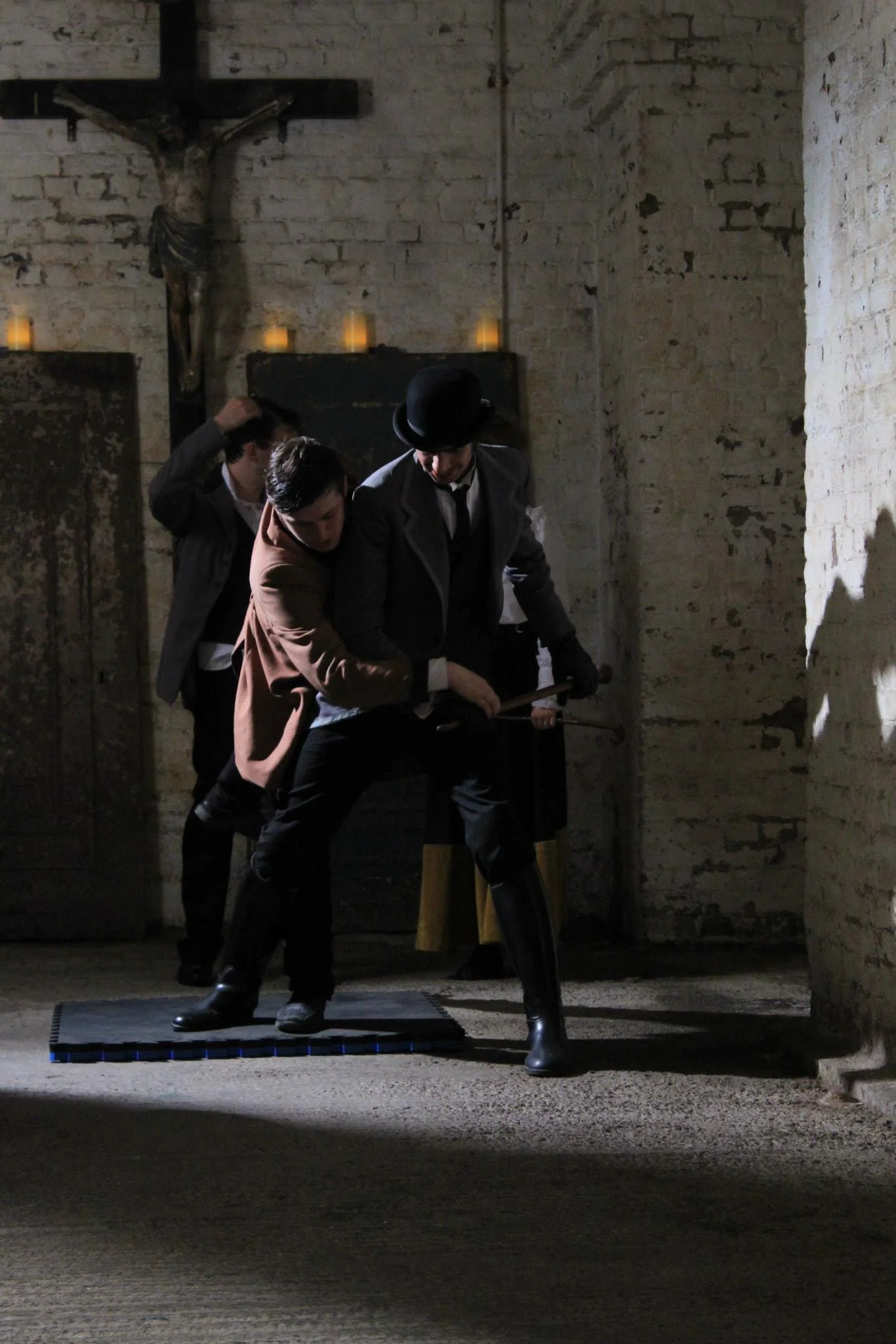 Three men in vintage clothing practicing sword fighting in a dimly lit room with brick walls, candles, and a crucifix on the wall.
