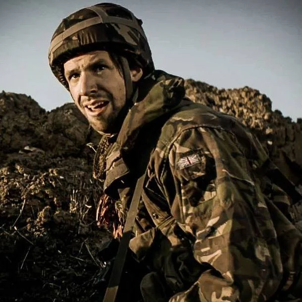 A soldier in camouflage uniform with a tactical helmet, standing outdoors in rocky terrain during daylight.