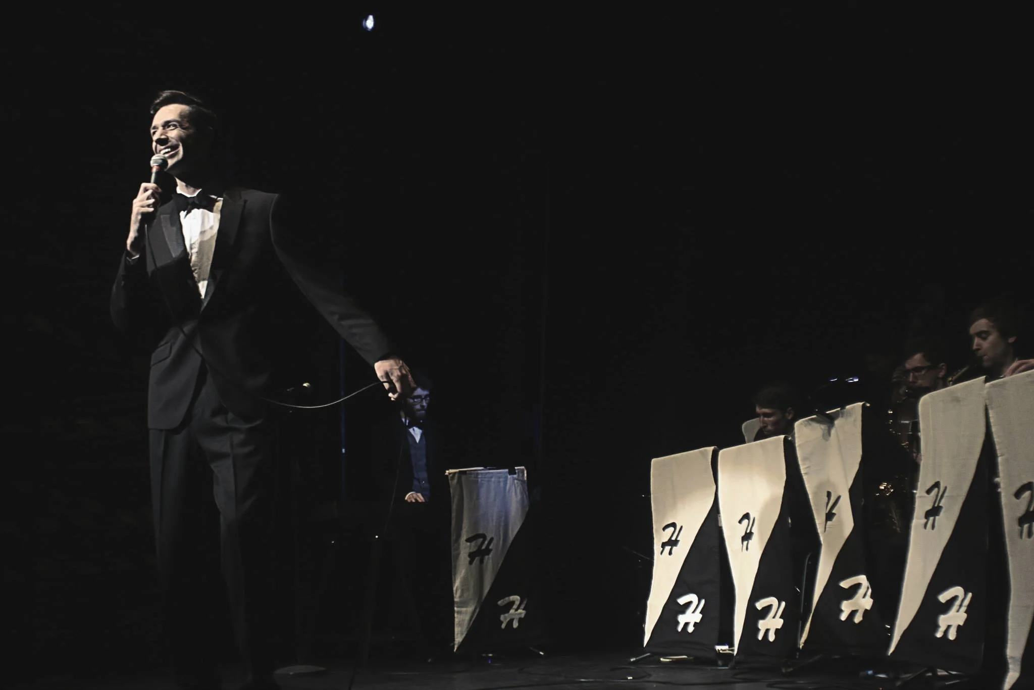 A man in a tuxedo holding a microphone, standing on a stage with a dark background. Several people are sitting in chairs along the stage, members of the band, behind music stands covered with black and white cloth marked "H H"