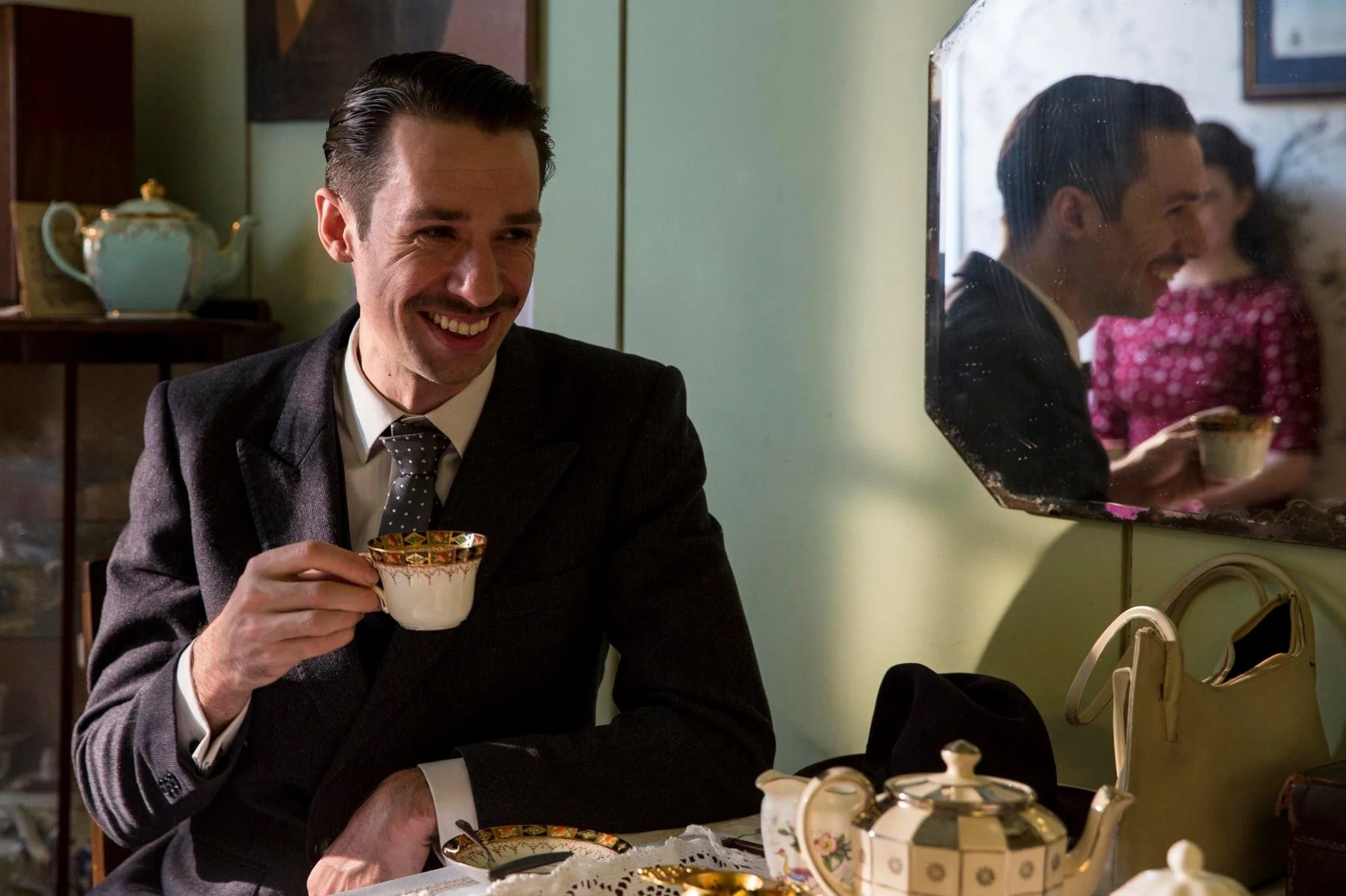 A man in a suit smiling and holding a cup at a tea table with a teapot, cup, and saucers in front of a mirror in a vintage-style room.