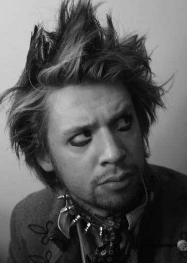 Black and white close-up of a young man with spiky, tousled hair, wearing a jacket and bandana, looking to the side with a serious expression.
