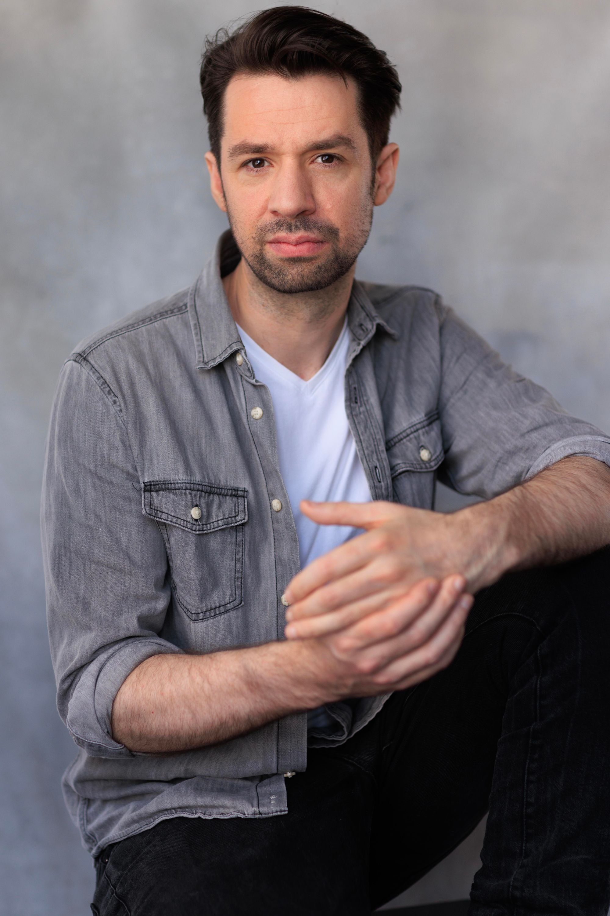 A man with dark hair and a beard wearing a gray denim shirt over a white T-shirt, sitting against a neutral gray background, looking into the camera with a serious expression.