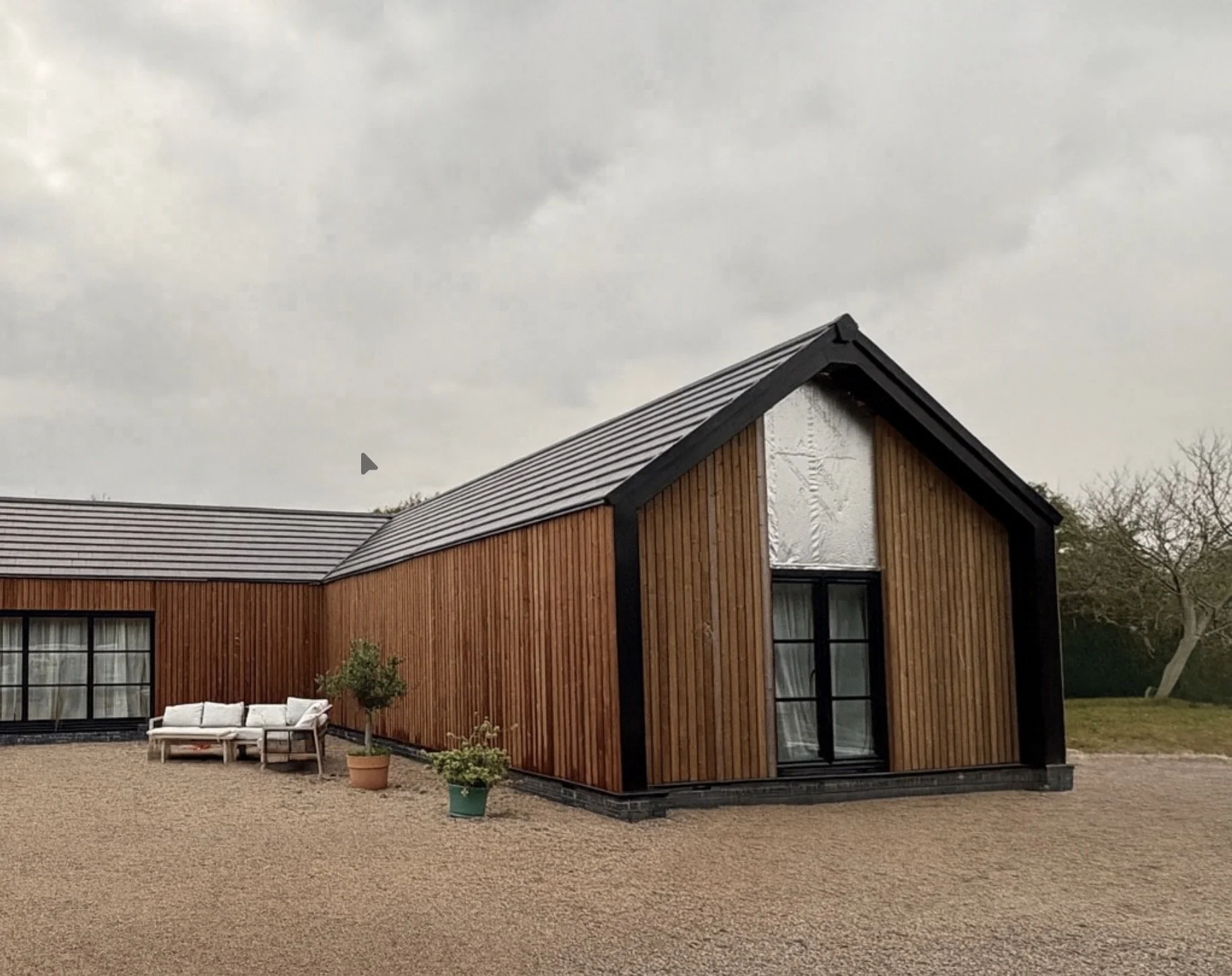 A modern house with wooden exterior walls, black trim, and a sloped metal roof. There are large windows, a white outdoor