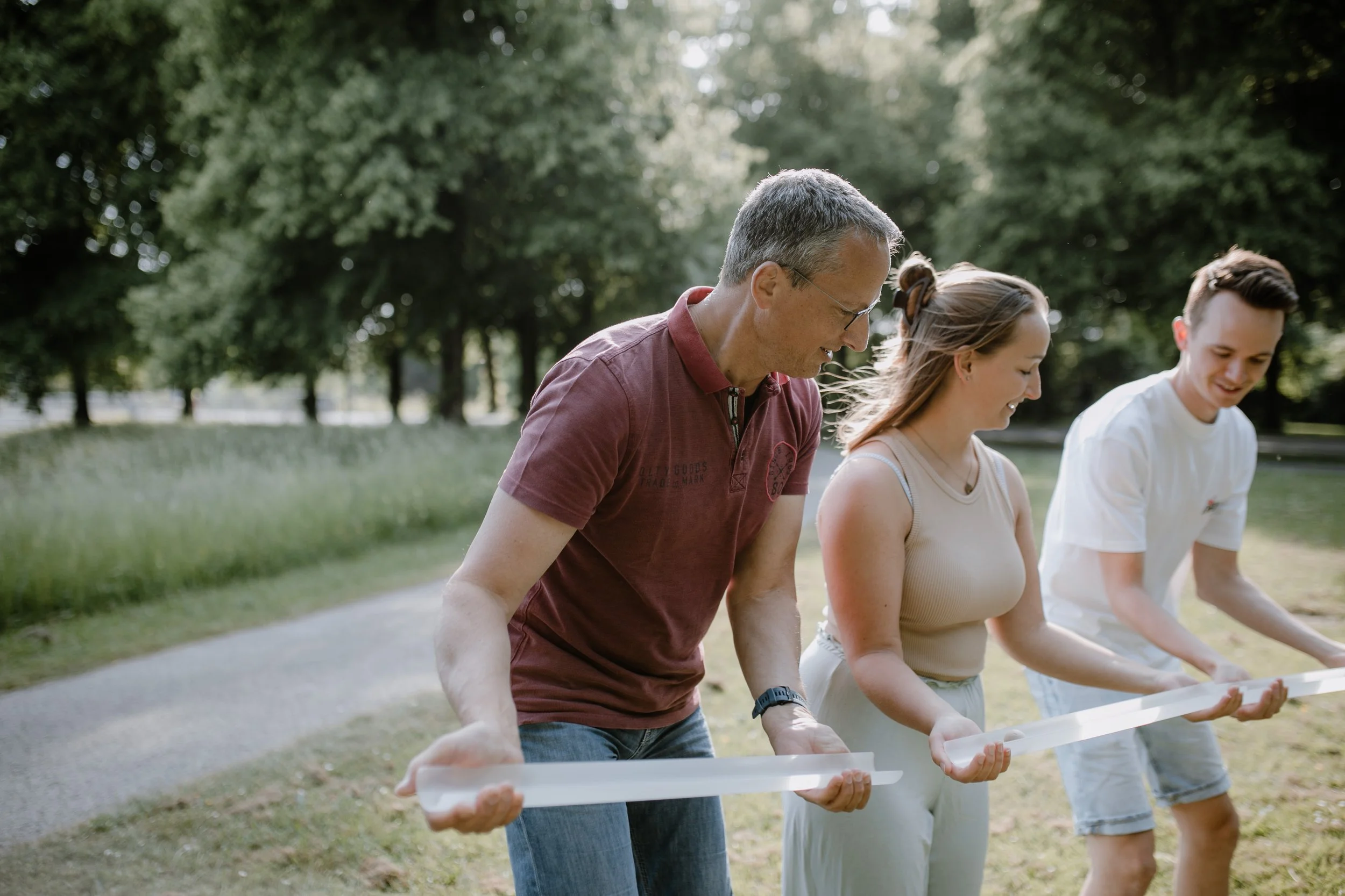 Drei Menschen spielen im Park mit einem langgestreckten Gegenstand, umgeben von Bäumen und grasbewachsenem Gebiet.