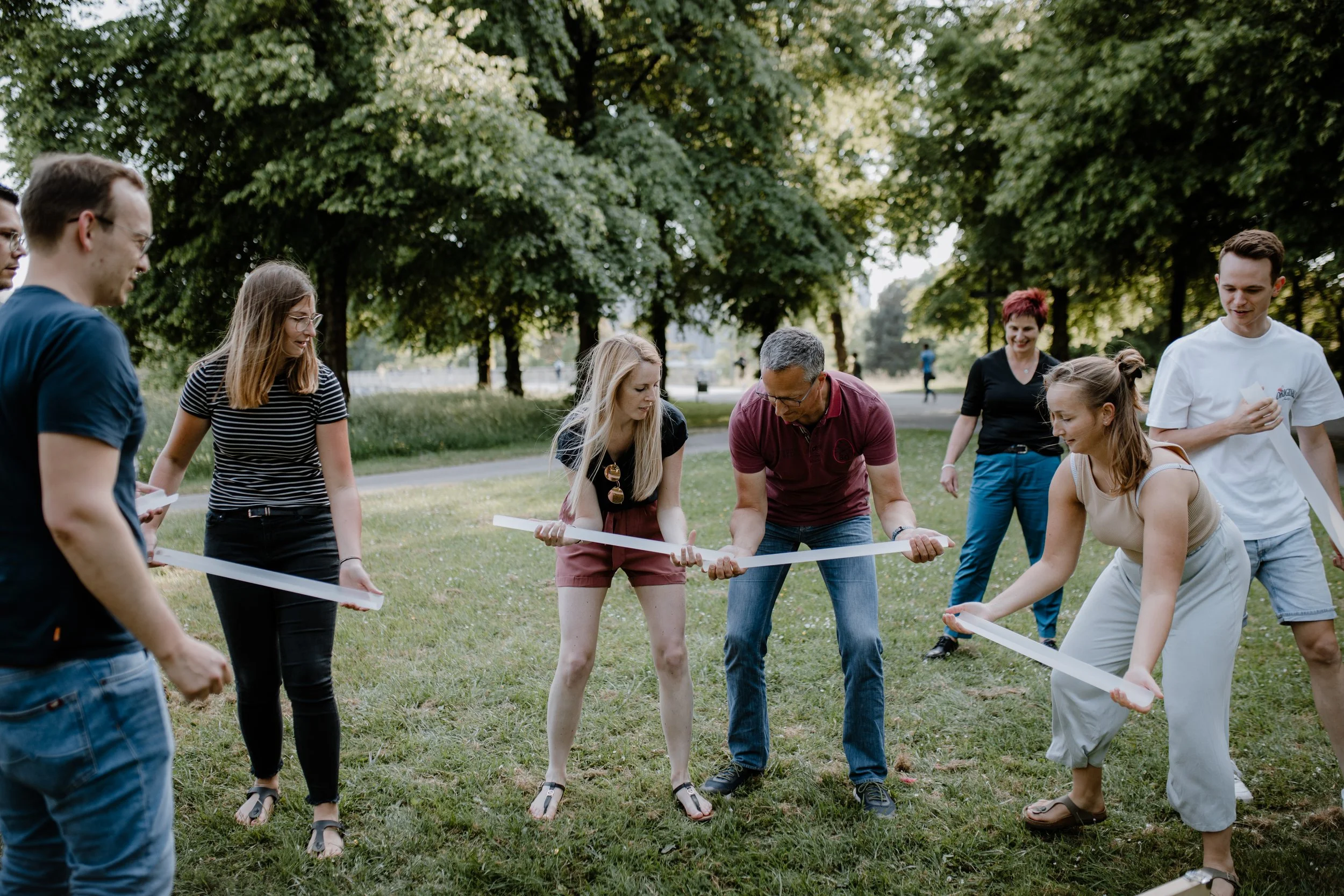 Gruppe von Menschen, die im Park an einem Team-Building-Workshop teilnehmen, bei dem sie mit weißen Reifen spielen.