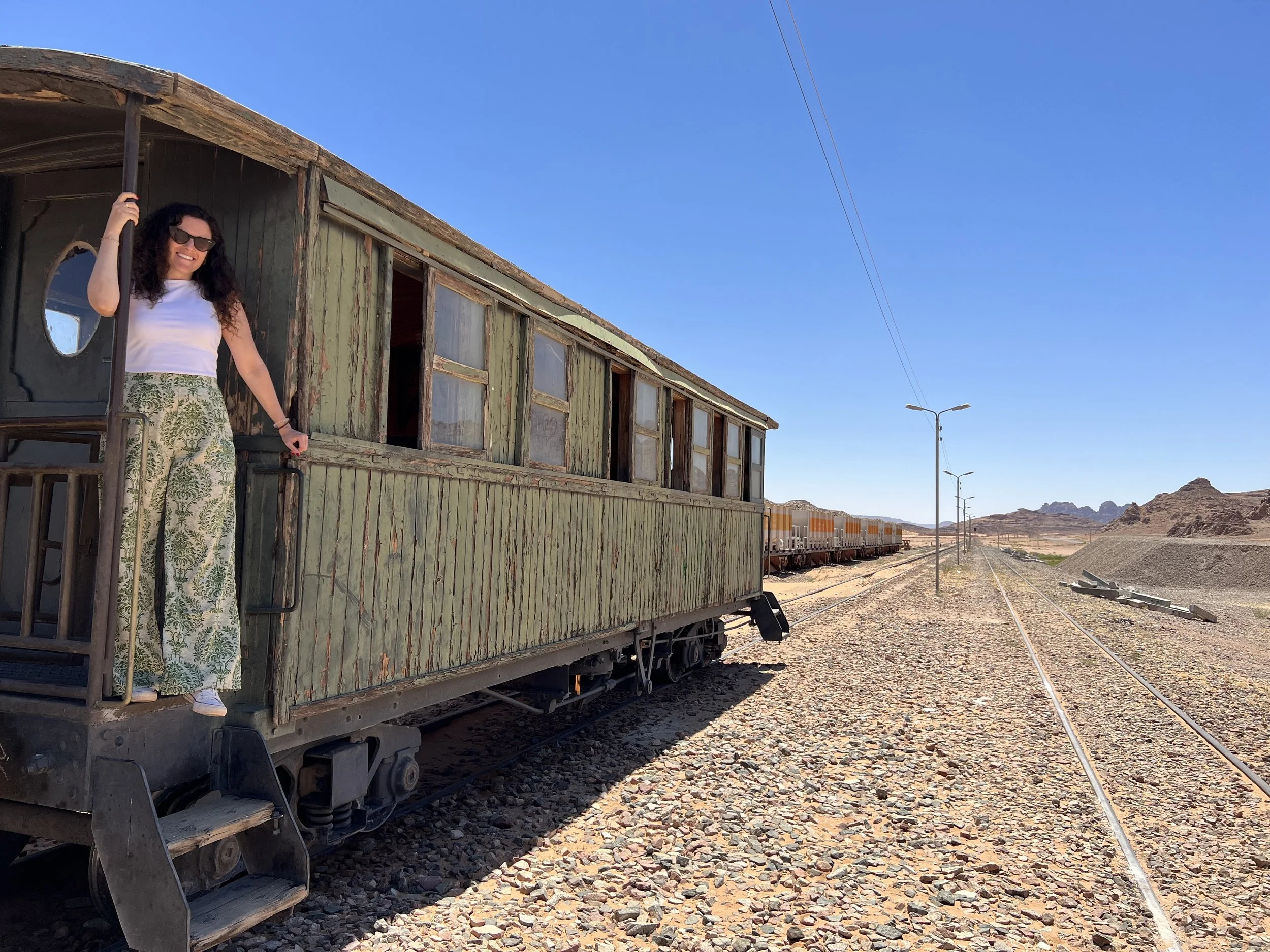 A woman with long curly hair wearing sunglasses, a white top, and patterned wide-leg pants, standing on the porch of an old weathered green train carriage in a desert landscape under a clear blue sky.