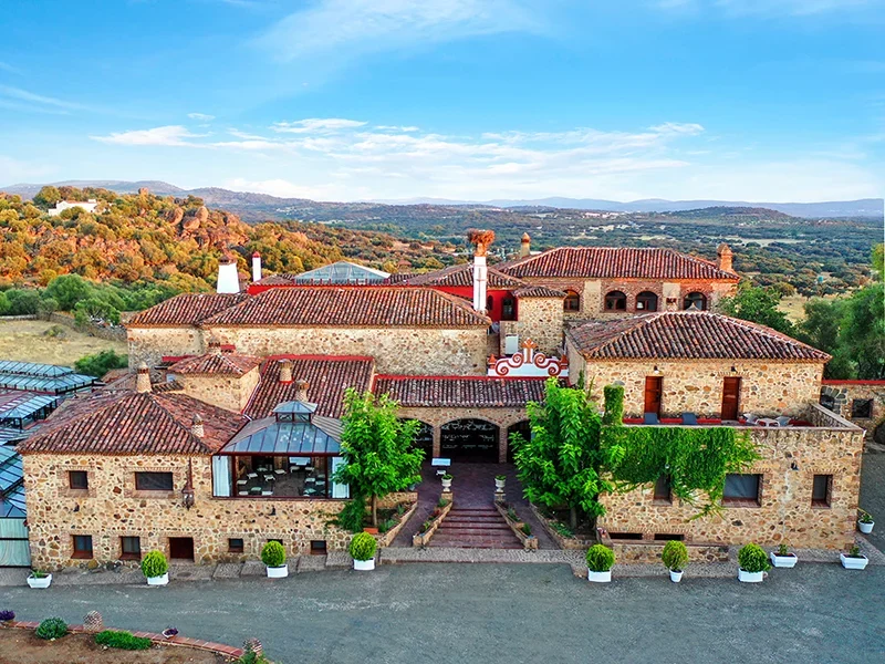 A large, rustic stone mansion with red-tiled roofs, surrounded by trees and rolling hills under a blue sky.