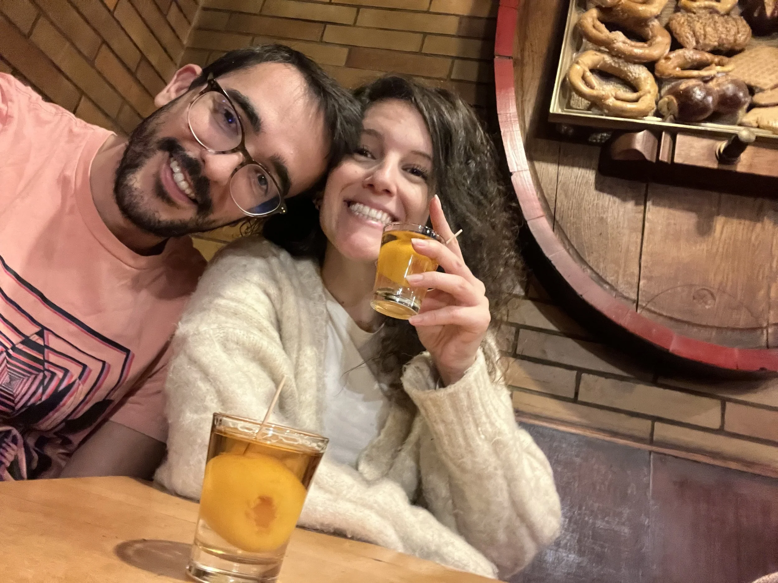 A man and a woman sitting at a wooden table in a restaurant, smiling for a selfie. The woman is holding a glass of beer, and there is a glass with a peach slice in front of her. The background features a brick wall and a large decorative wooden barrel with various pretzels and sausages on display.