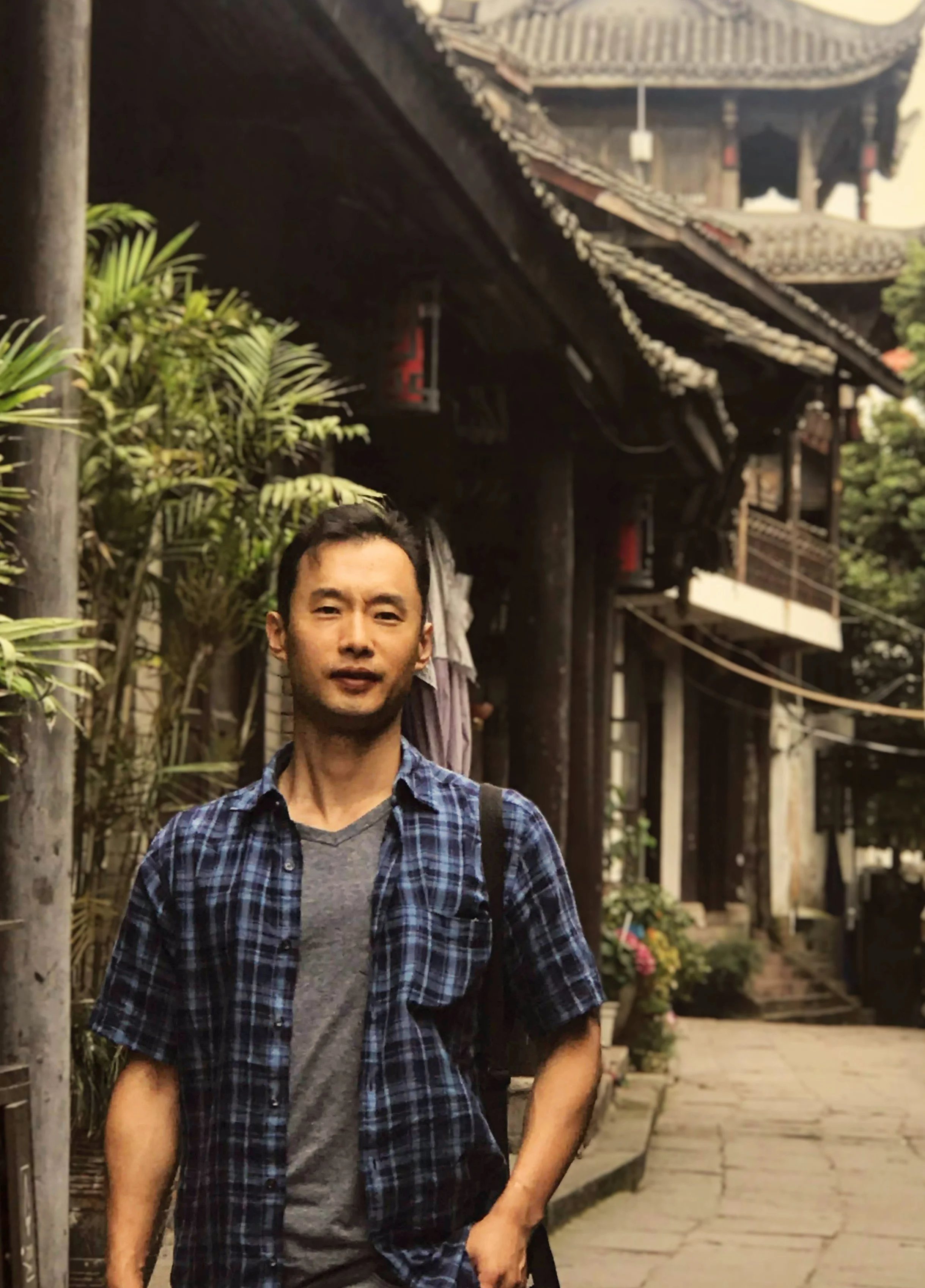 A man standing outdoors on a stone pathway near traditional Asian-style wooden buildings with curved rooflines, greenery, and potted plants.