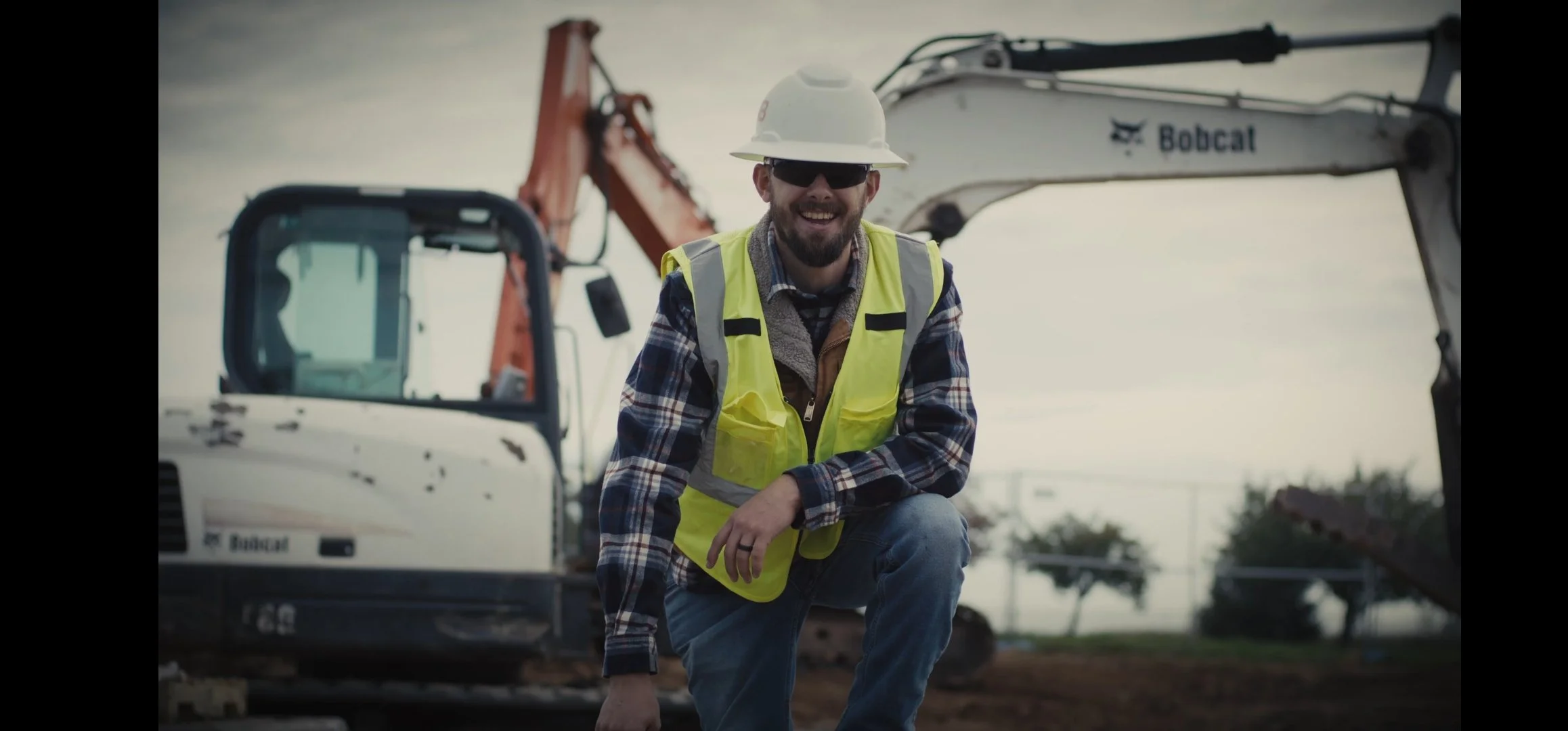 A man in a yellow safety vest, hard hat, and sunglasses kneeling in front of construction equipment, smiling on a construction site with an overcast sky.