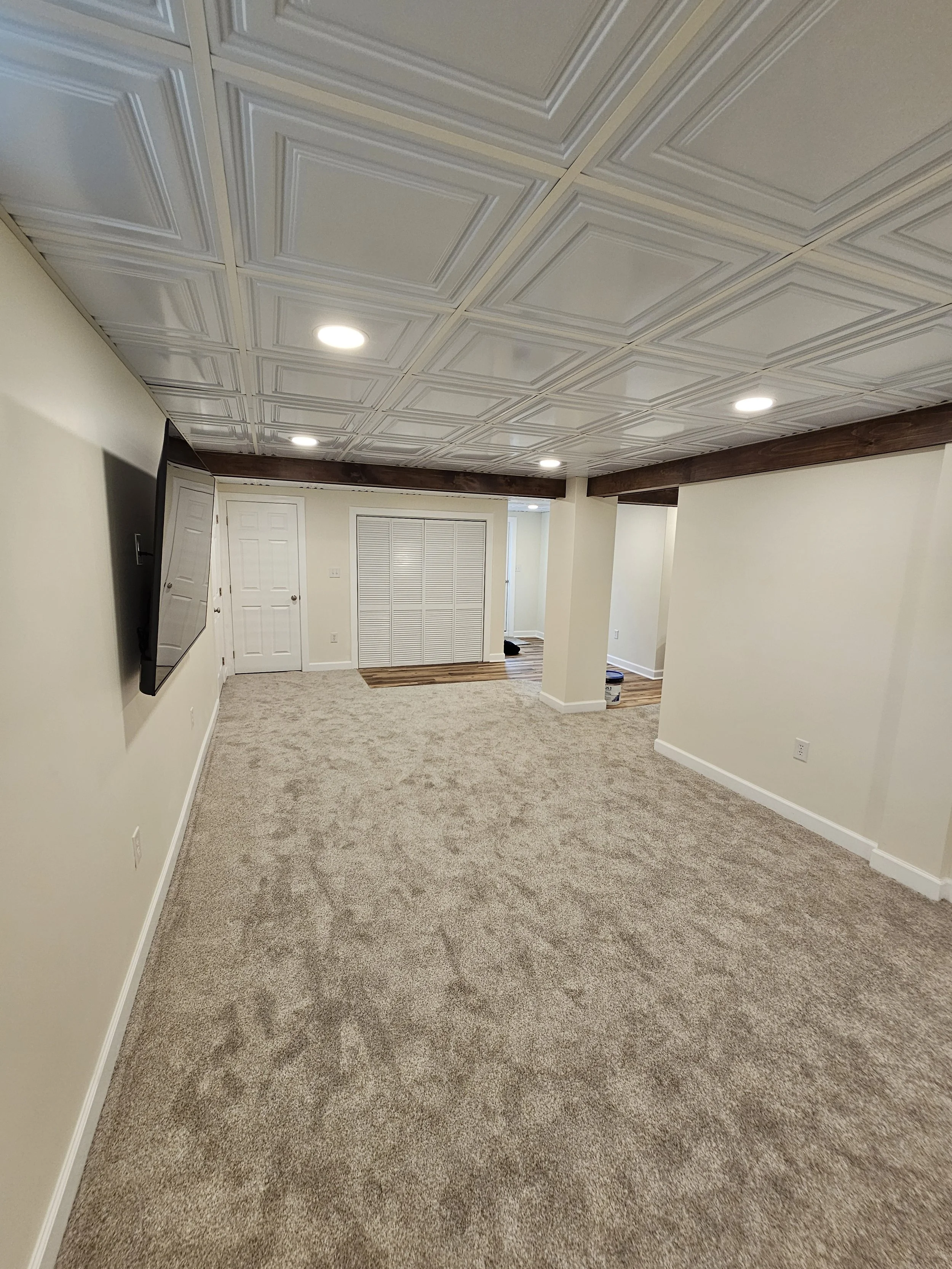 Empty basement room with beige carpet, white walls, a wall-mounted flat-screen TV, wood beams, and ceiling with decorative tiles and recessed lighting.