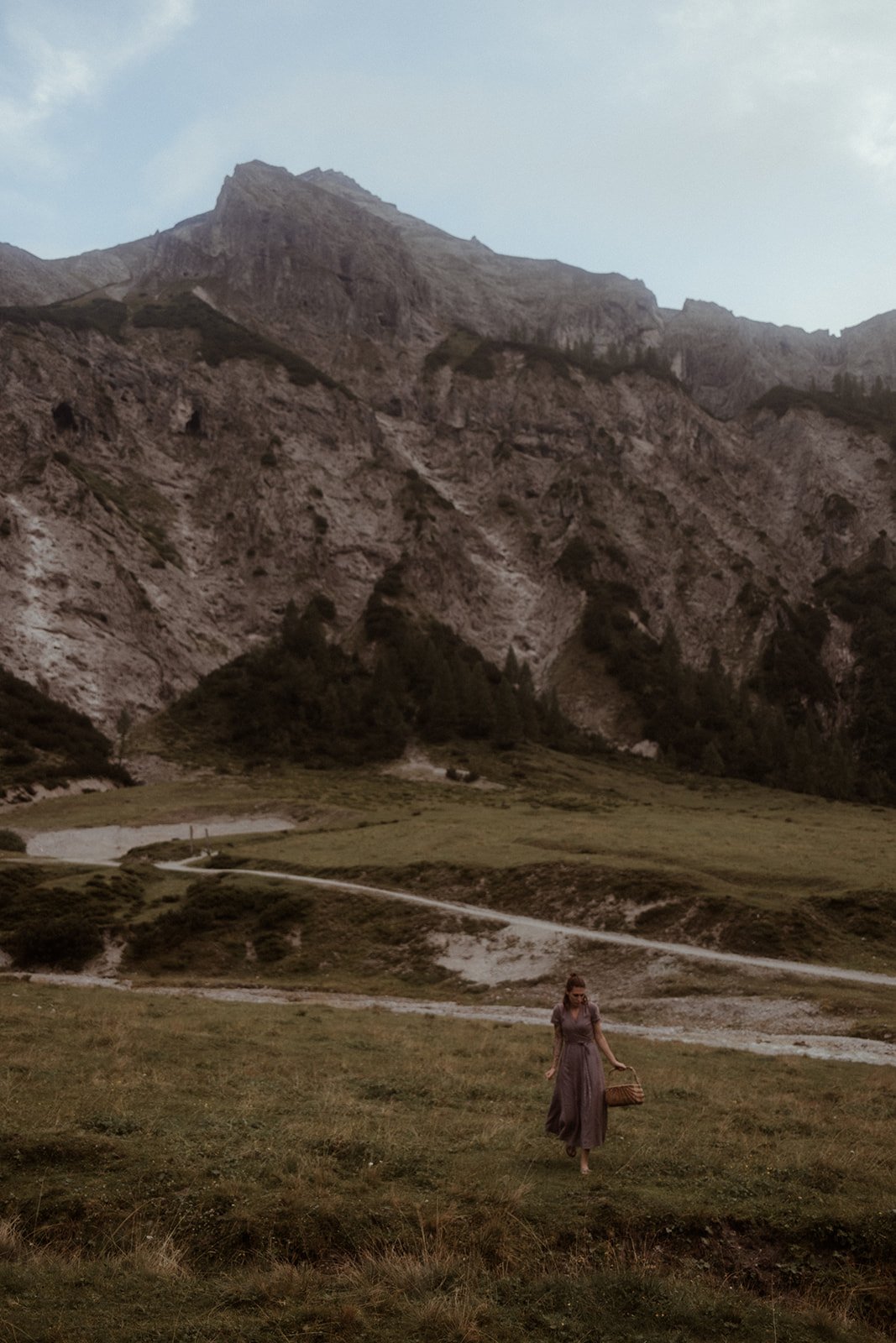 Eine Frau in einem langen, grauen Kleid trägt einen Korb und spaziert durch eine grüne Wiesenlandschaft vor hohen, felsigen Bergen.