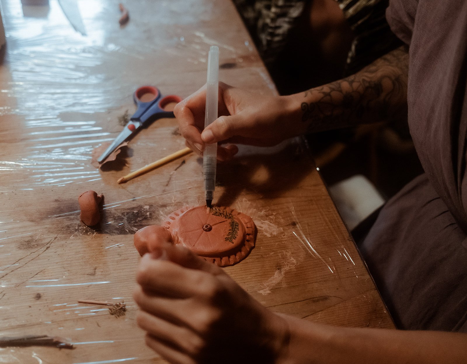 Eine Person dekoriere eine bemalte Backform mit einem Filzstift. Auf dem Tisch liegen Schere, Holzstäbchen und Ton. Der Tisch ist mit Klarsichtfolie bedeckt.