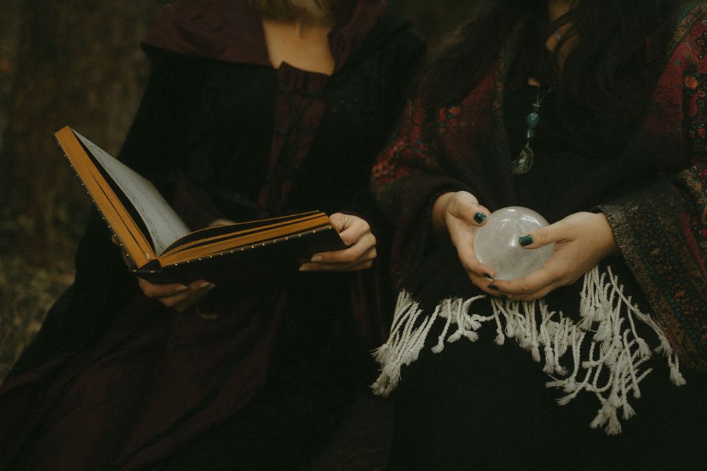 Zwei Frauen sitzen nebeneinander, eine liest in einem großen offenen Buch, die andere hält eine gläserne Kristallkugel. Beide tragen dunkle, bunte Kleidung mit Fransen und Accessoires.