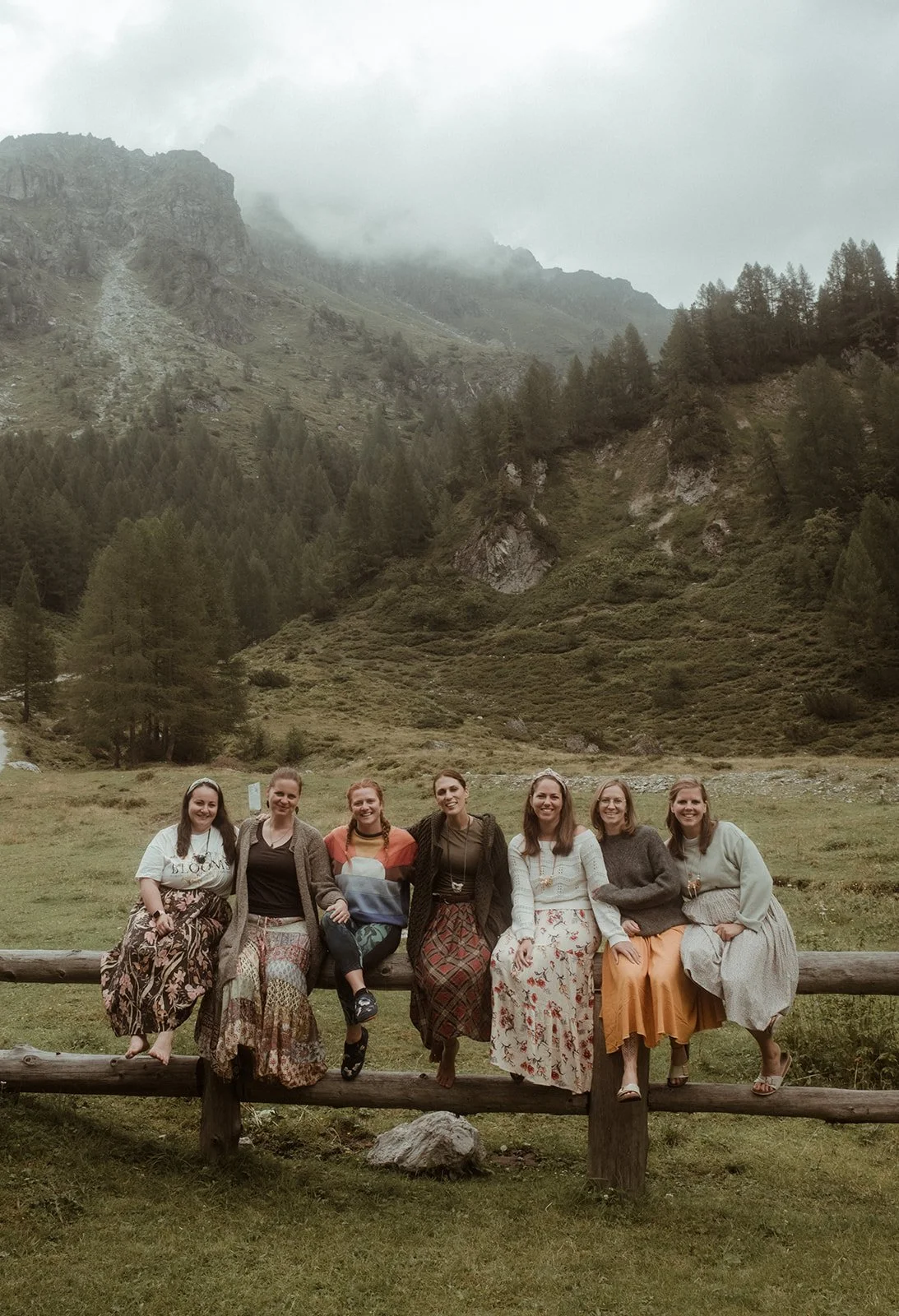 Sieben Frauen sitzen auf einem Holzgeländer vor einer malerischen Bergkulisse, umgeben von grünen Hügellandschaften und bewölktem Himmel.
