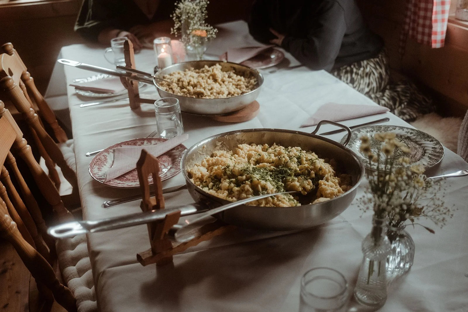 Ein festlich gedeckter Esstisch mit zwei großen Servierpfannen voller Kartoffelsalat, umgeben von Geschirr, Gläsern und Blumen.