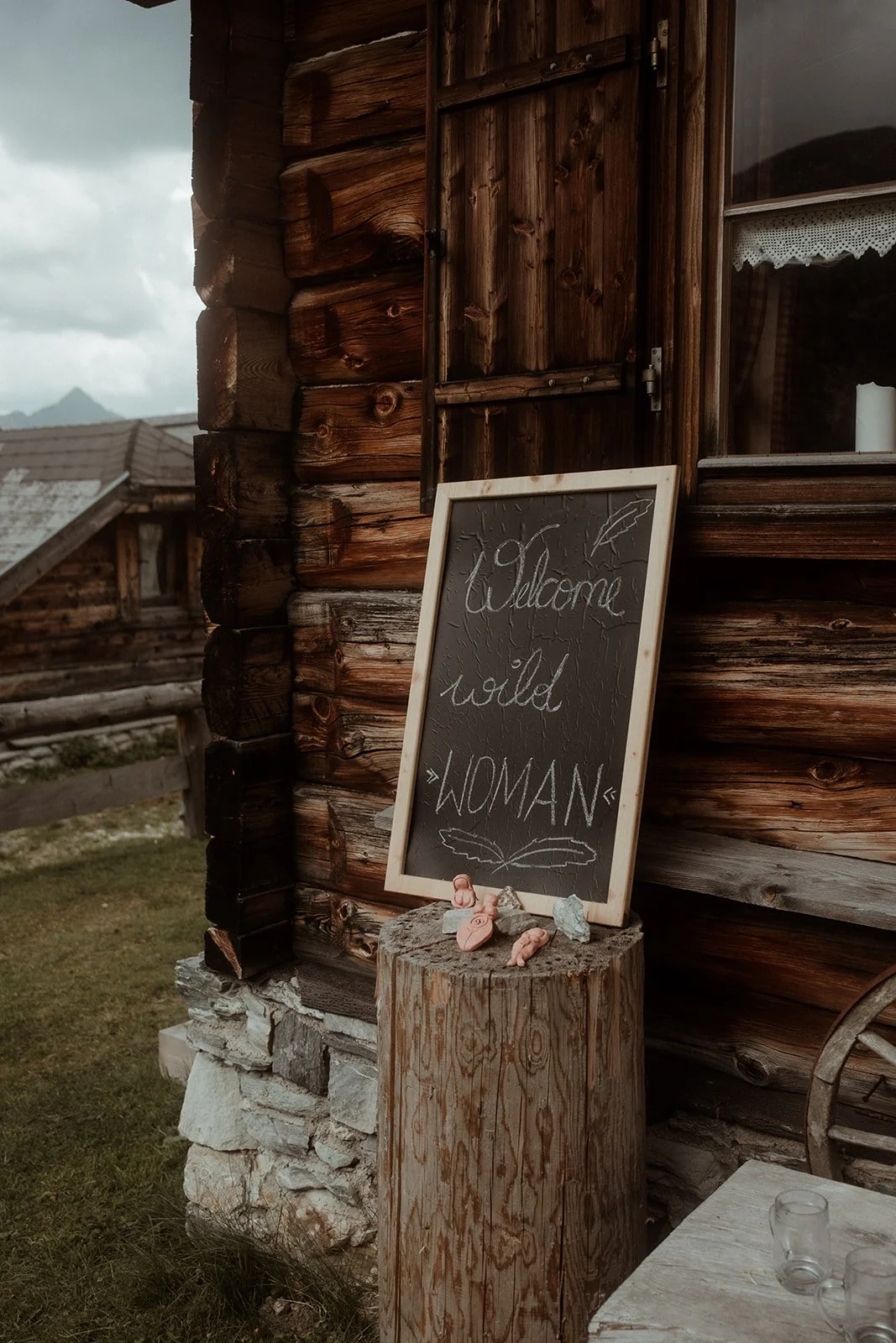 Ein Holzwand mit Fenster und einem schwarzen Wandtafel mit weißen Schriftzug 'Welcome cold WOMAN' in einer rustikalen Atmosphäre, umgeben von kleinen Dekorationsartikeln auf einem Baumstamm