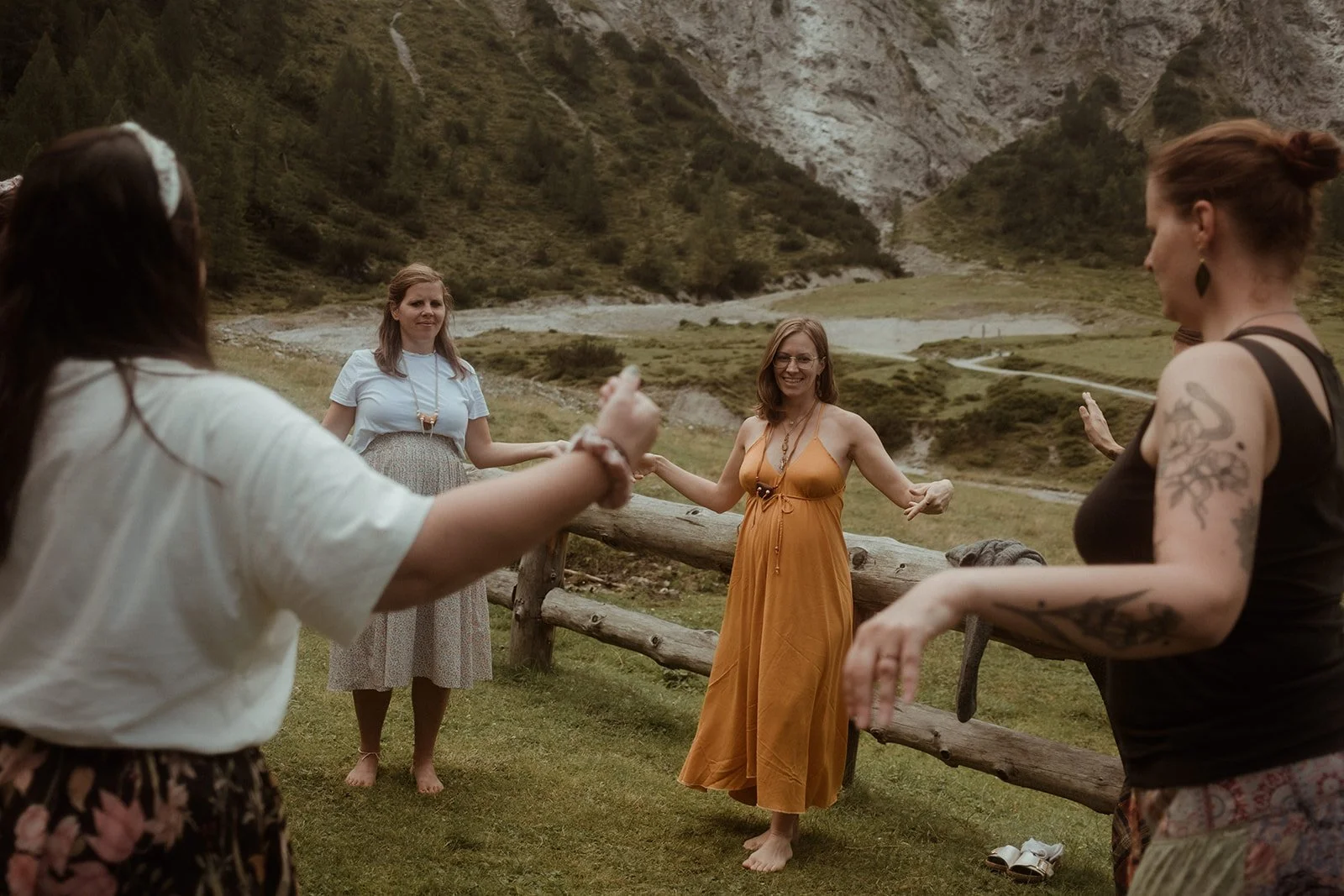 Frau in einem orangefarbenen Kleid tanzt mit anderen Frauen in einer natürlichen Landschaft mit Hügeln und einem Bach im Hintergrund.