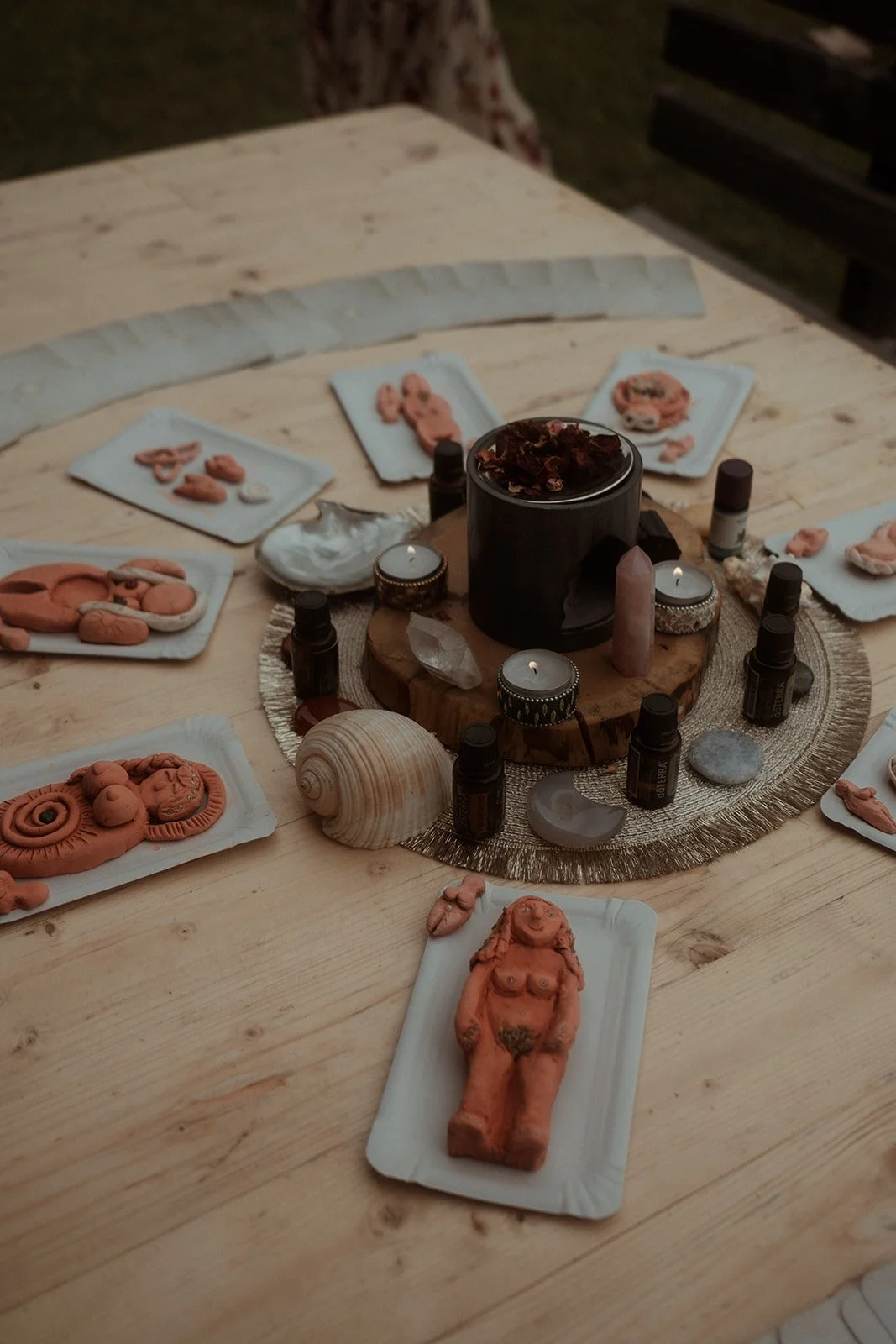 Ein Tisch mit einer runden goldfarbenen Unterlage, auf der mehrere Räucherküchen, Teelichter, einige kleine Fläschchen und Kristalle platziert sind. Darauf liegen Tonfiguren und Gegenstände, die wie geformte Figürchen, Muscheln und Steine aussehen.