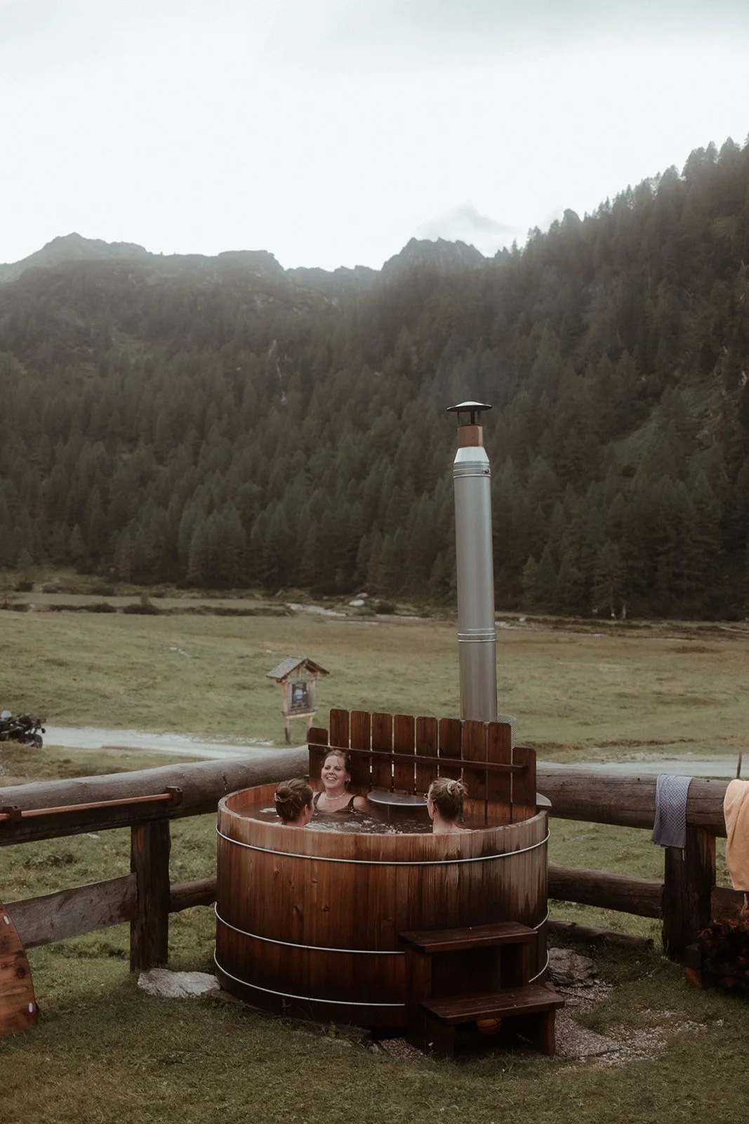 Drei Frauen sitzen in einem runden Holzbad im Freien auf einer Wiese, umgeben von Bergen und Wäldern, während eine Person im Hintergrund im Wasser ist.