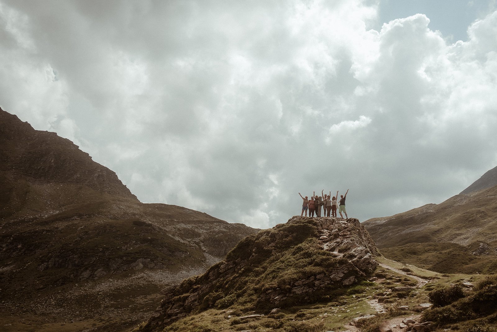 Gruppe von Menschen auf einem Felsen in einer Berglandschaft bei bewölktem Himmel, die jubeln und die Arme in die Luft strecken.