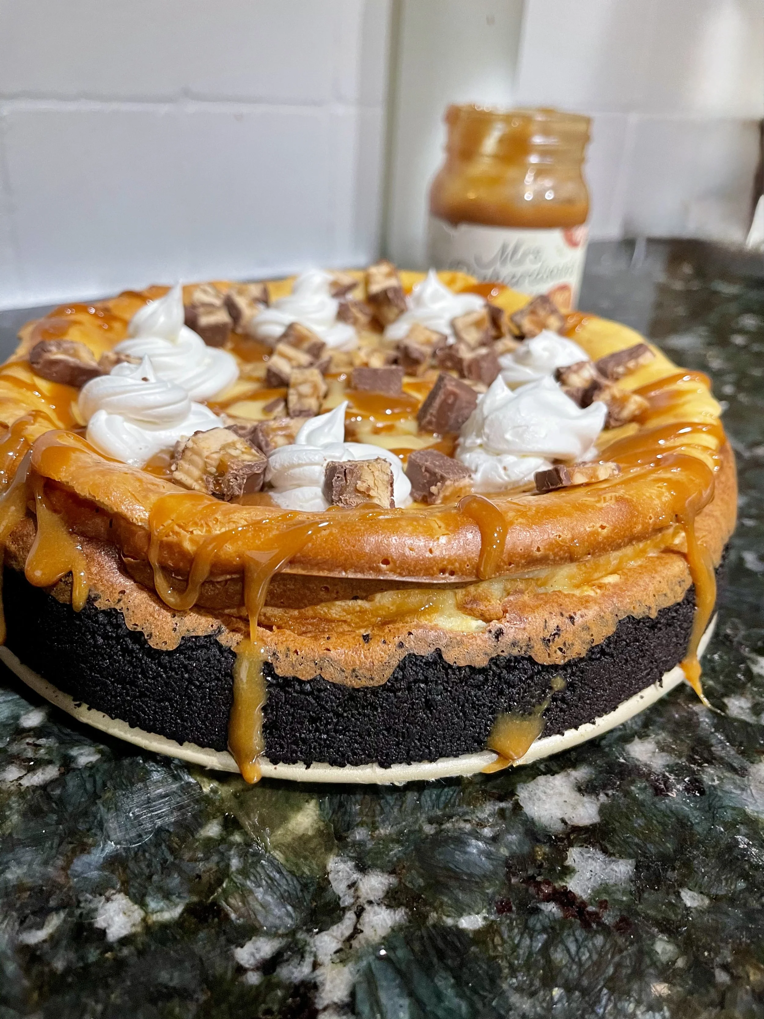 A cheesecake with a chocolate crust, topped with caramel sauce, whipped cream, and chopped Reese's Peanut Butter Cups.