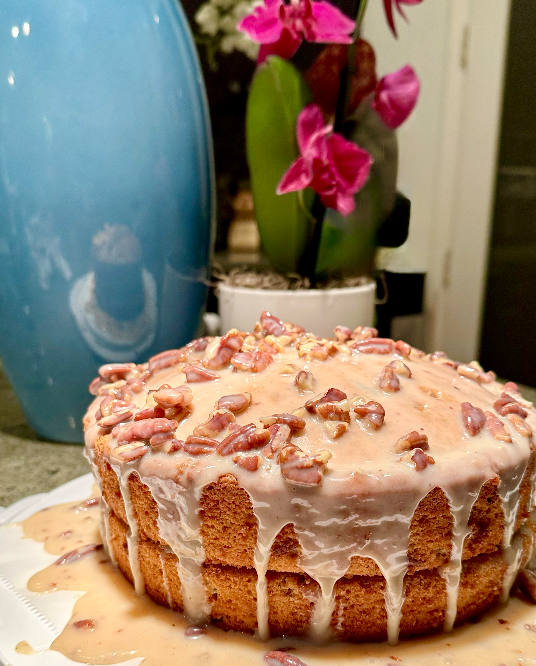 A round cake with caramel glaze and chopped pecans on top, with some glaze dripping down the sides, placed on a white cake stand.