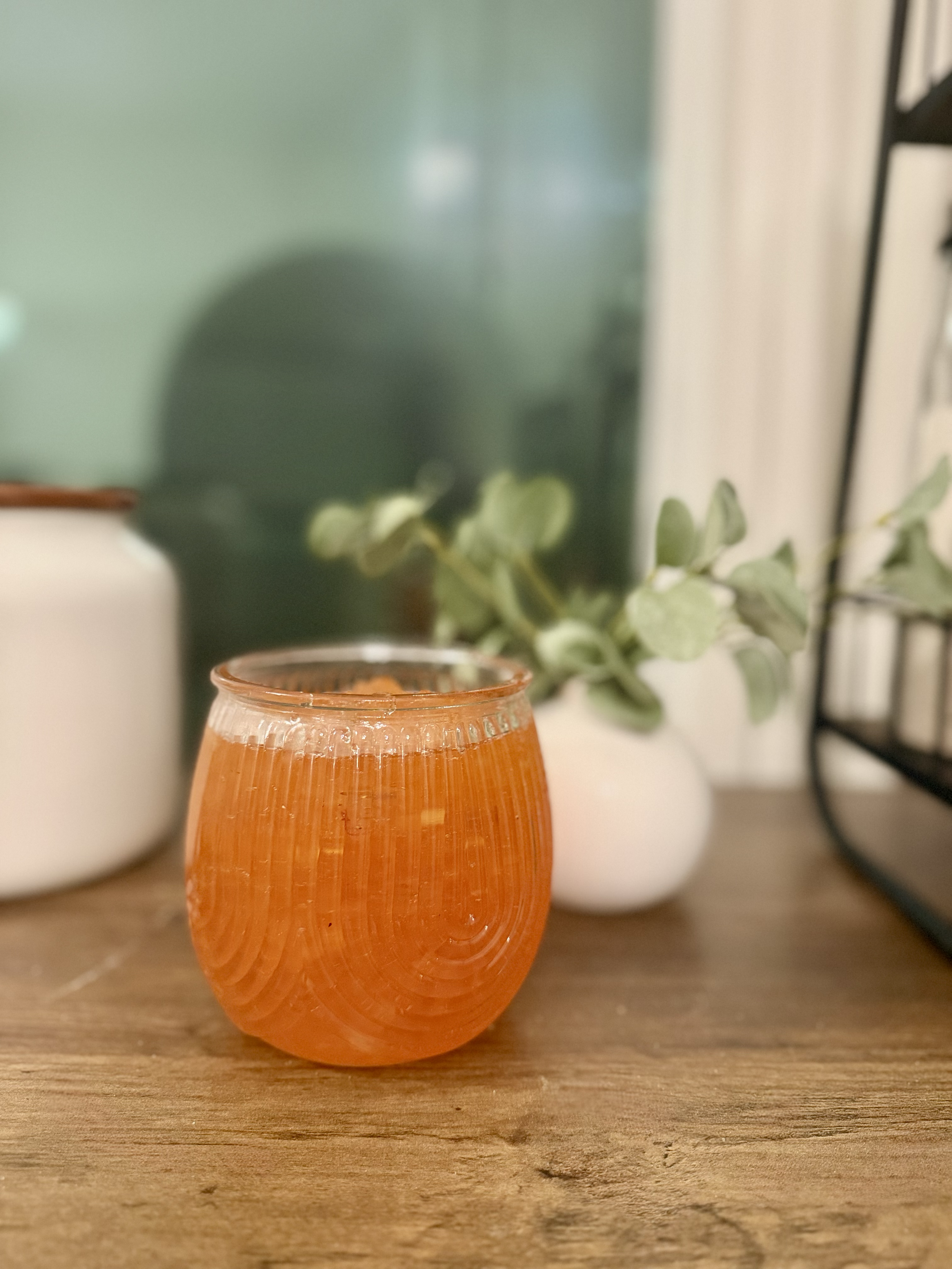 Strawberry apple jam in a decorative jar, on a wooden surface with white vases and green leaves in the background.