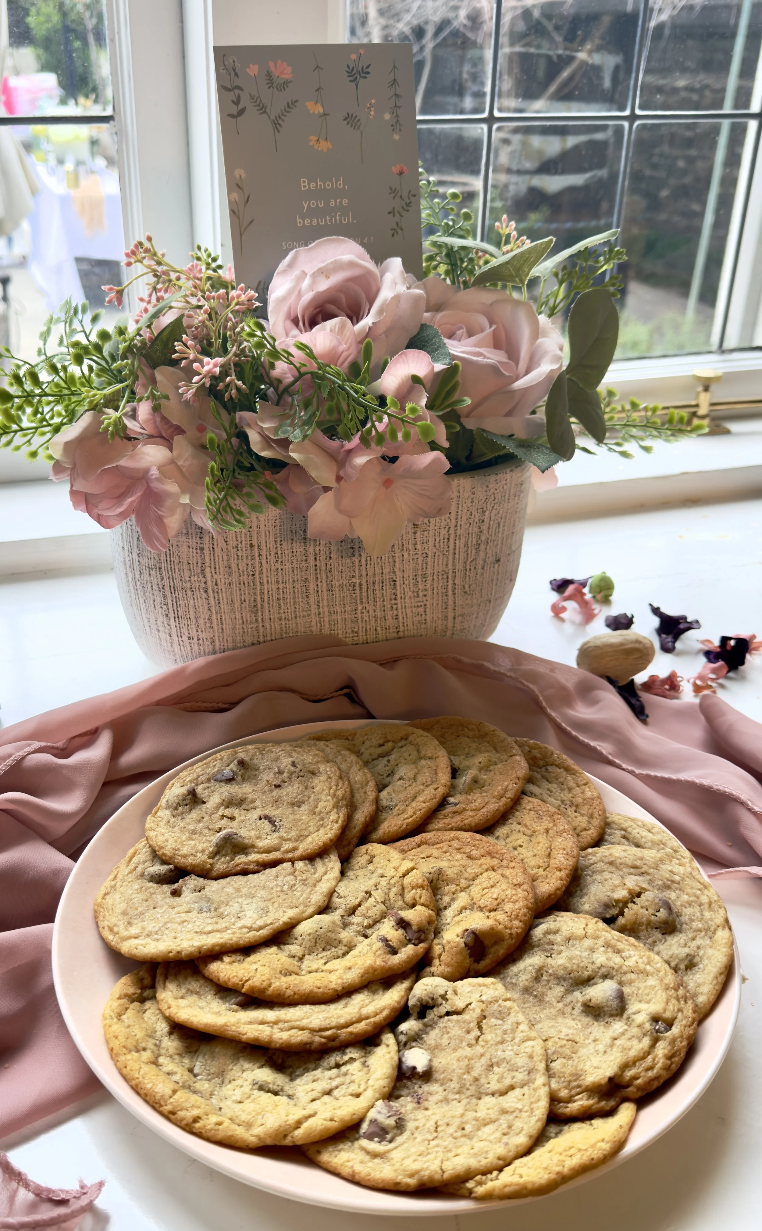 A plate of chocolate chip cookies on a pink cloth at a window, with a flower arrangement and a card with the words 'Behold, you are beautiful.' in the background.