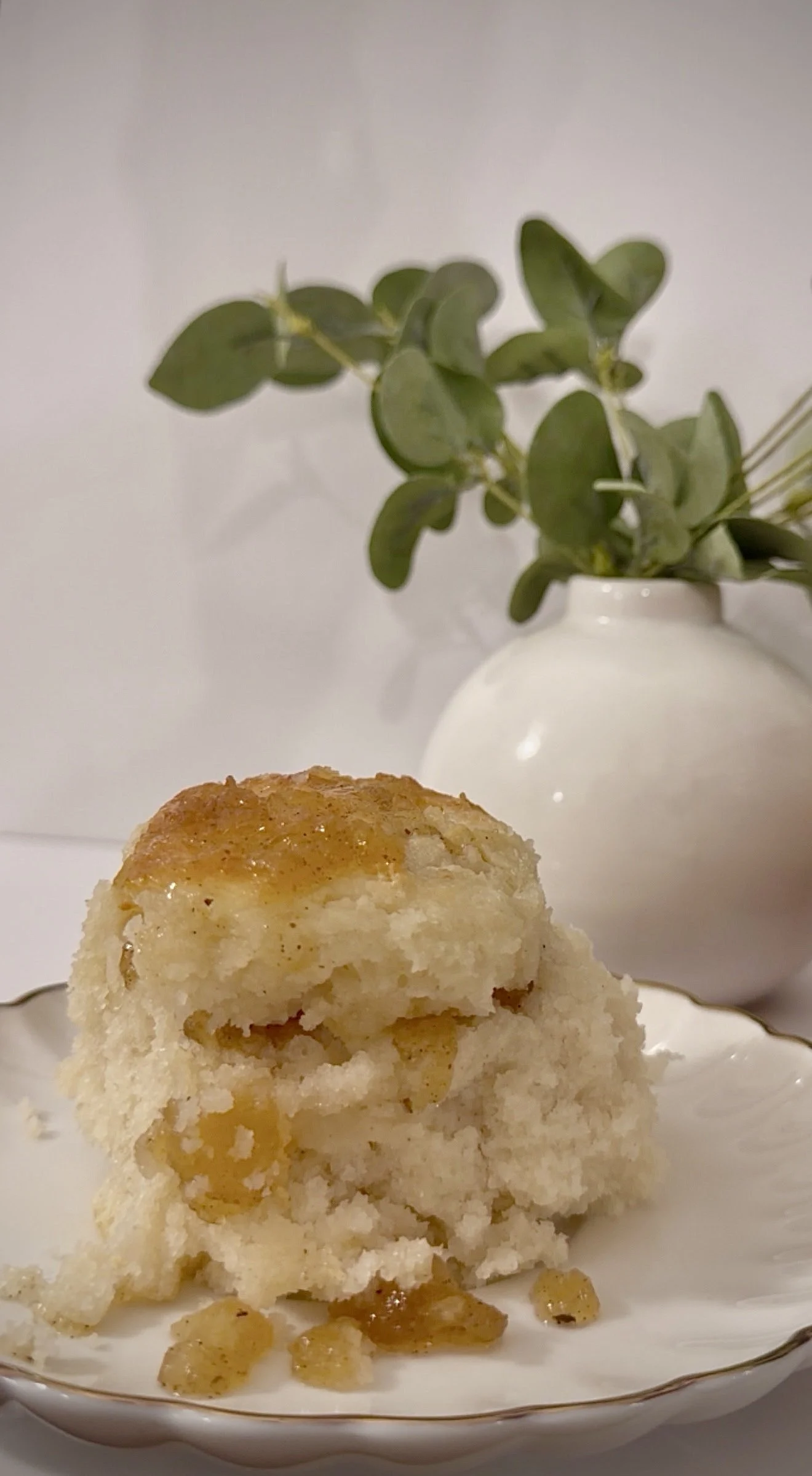A serving of buttermilk biscuit and apple pie jam with visible apple chunks and a golden-brown crust, on a white plate with a gold rim, placed on a white surface. In the background, there's a white vase with green leaves and small branches.