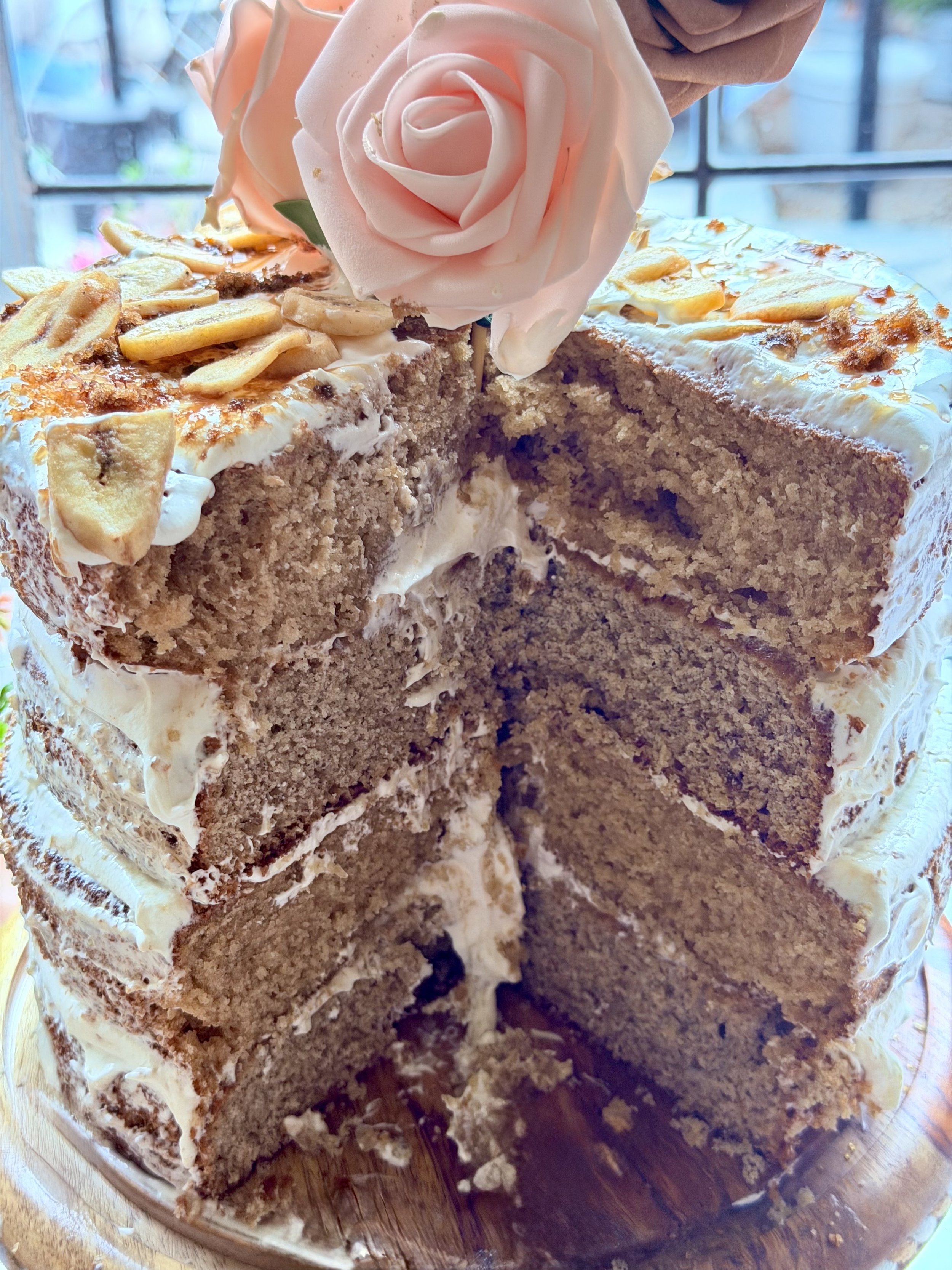 A layered Banana Fosters cake with cream frosting, topped with sliced bananas and a pink rose, with a person cutting a slice.