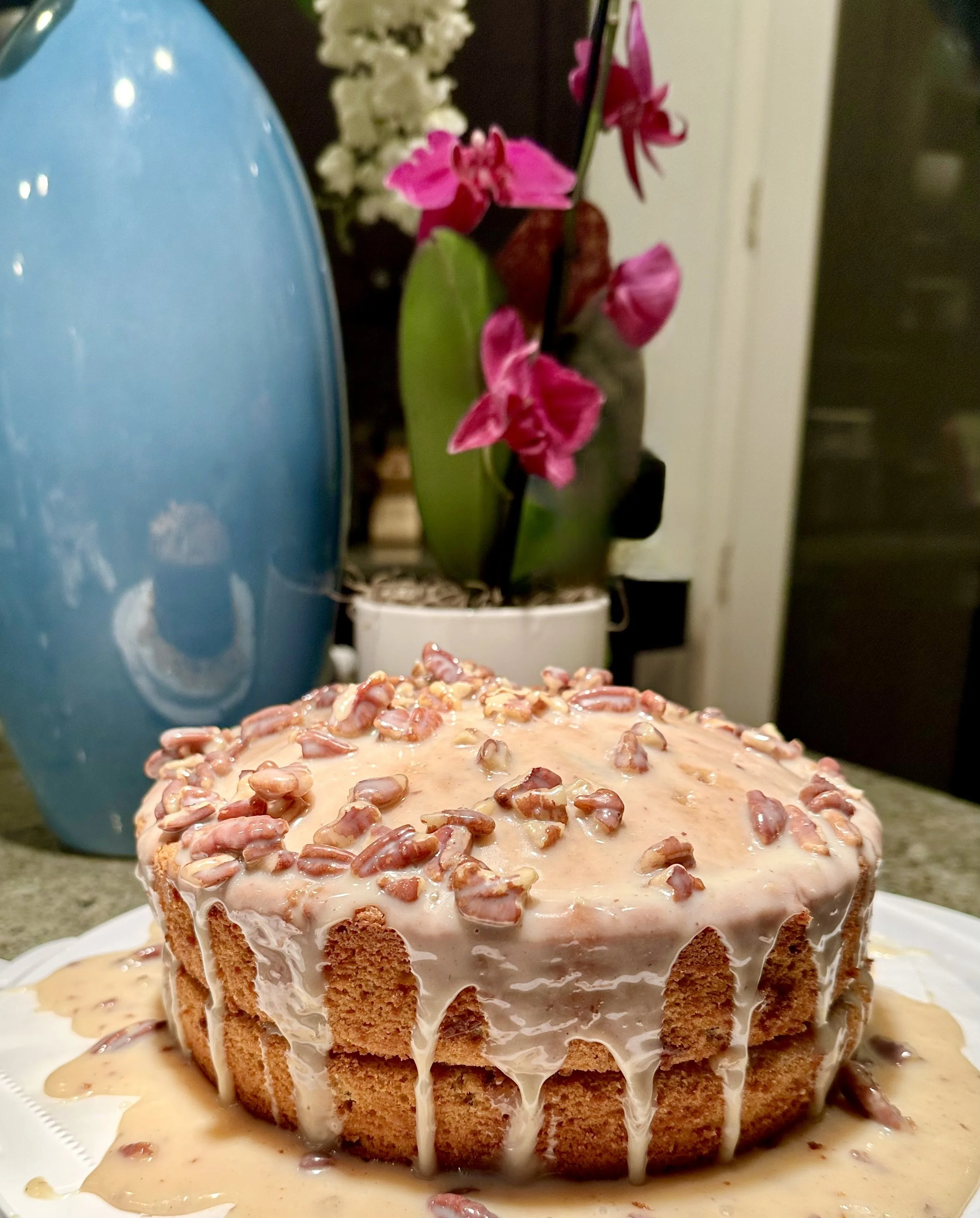 A round, layered cake with caramel-colored icing and chopped nuts on top, with caramel sauce dripping down the sides, placed on a white plate on a green countertop. In the background, there is a large blue vase, a potted pink orchid, and a white flower arrangement.