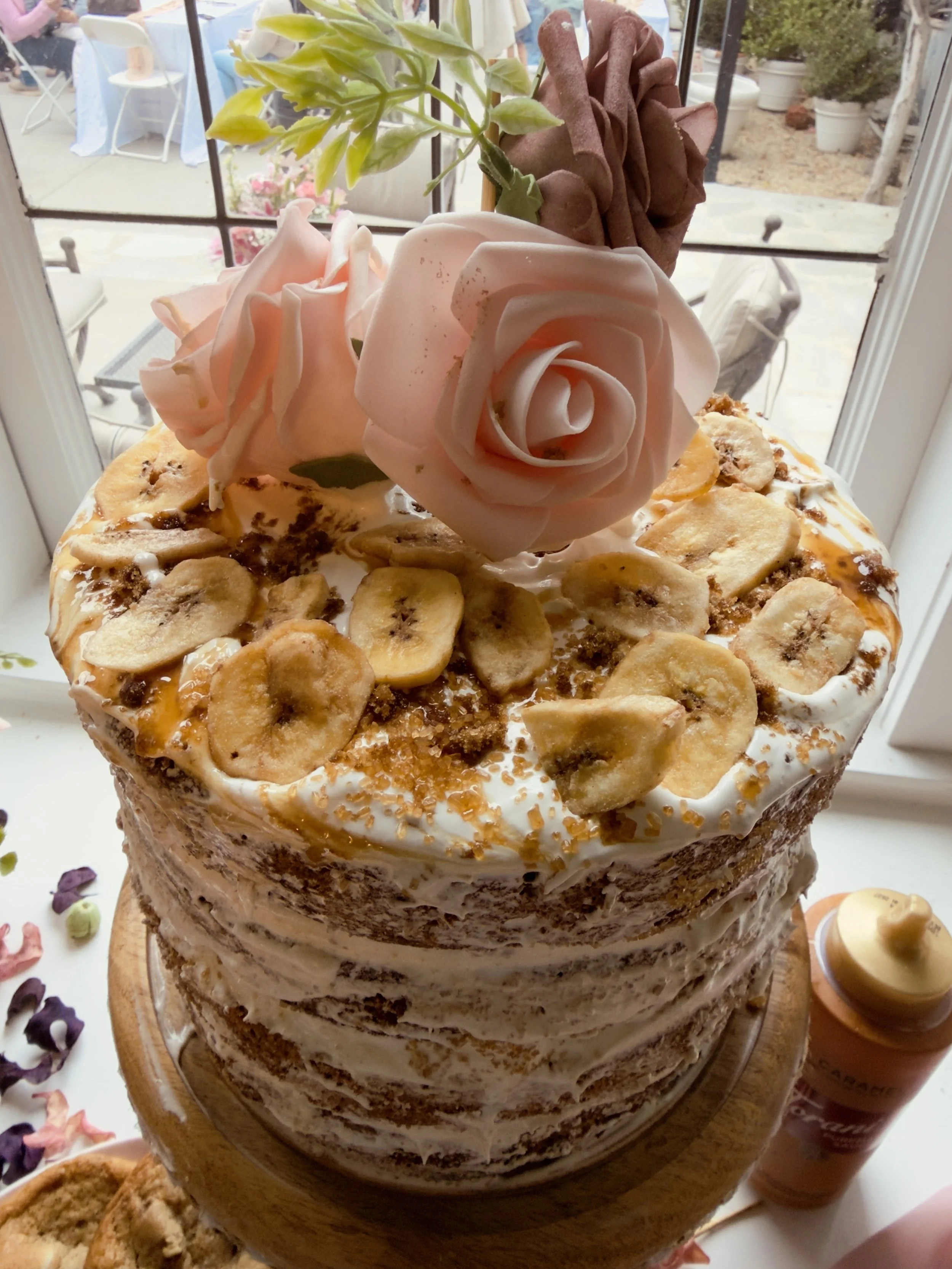 A layered cake decorated with banana slices, caramel drizzle, pink and brown roses, and foliage on top, placed on a wooden stand near a window.