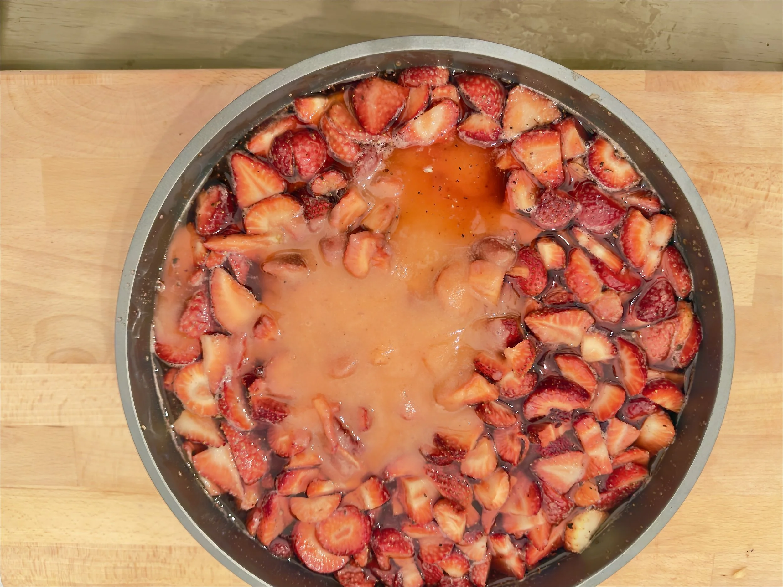 A round metal pan filled with halved strawberries  in sugar and lemon juice, on a wooden surface.