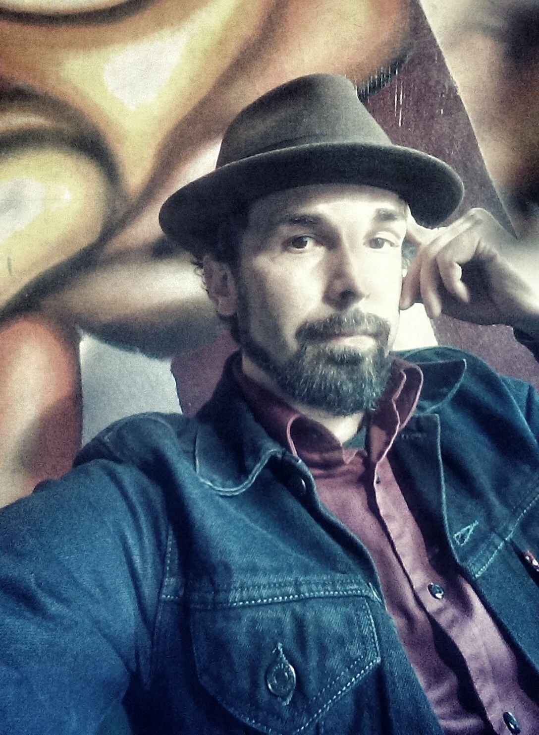A man with a beard and mustache wearing a fedora hat, a denim jacket, and a maroon shirt, posing with his hand resting on his temple in front of a mural or large photo of a close-up of a person's face.