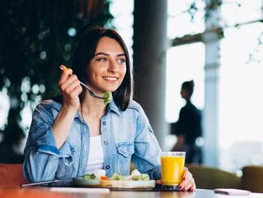 A young woman sitting at a table eating breakfast with a glass of orange juice, smiling, in a bright cafe with large windows and a blurred person in the background.