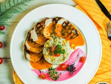 Chicken breast slices with grilled marks, served with a portion of rice topped with vegetables and a cherry, and a side of pink sauce garnished with herbs and cherry tomatoes on a white plate.