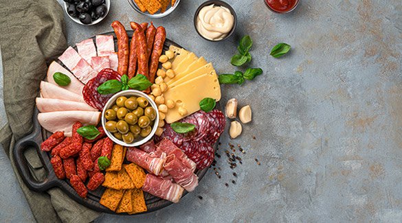 Variety of sliced meats, cheeses, and snack foods on a charcuterie board with olives, crackers, and sauces.