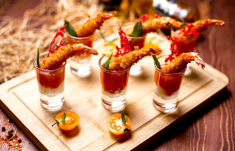 Six shot glasses filled with shrimp cocktail, garnished with green leaves, placed on a wooden serving tray, with cherry tomatoes and a bottle in the background.