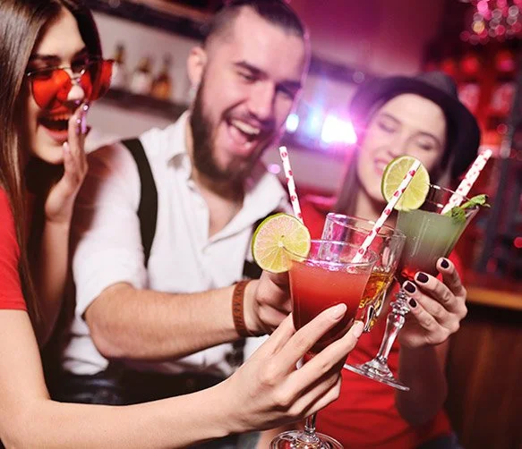 Three people, two women and one man, are smiling and holding colorful cocktails with lime slices and striped straws, celebrating together in a lively bar or club setting.