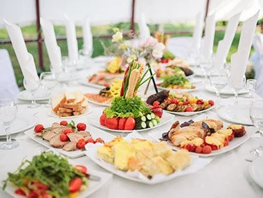 A table set for a meal with a variety of dishes, salads, and a centerpiece on a white tablecloth in a bright outdoor setting.